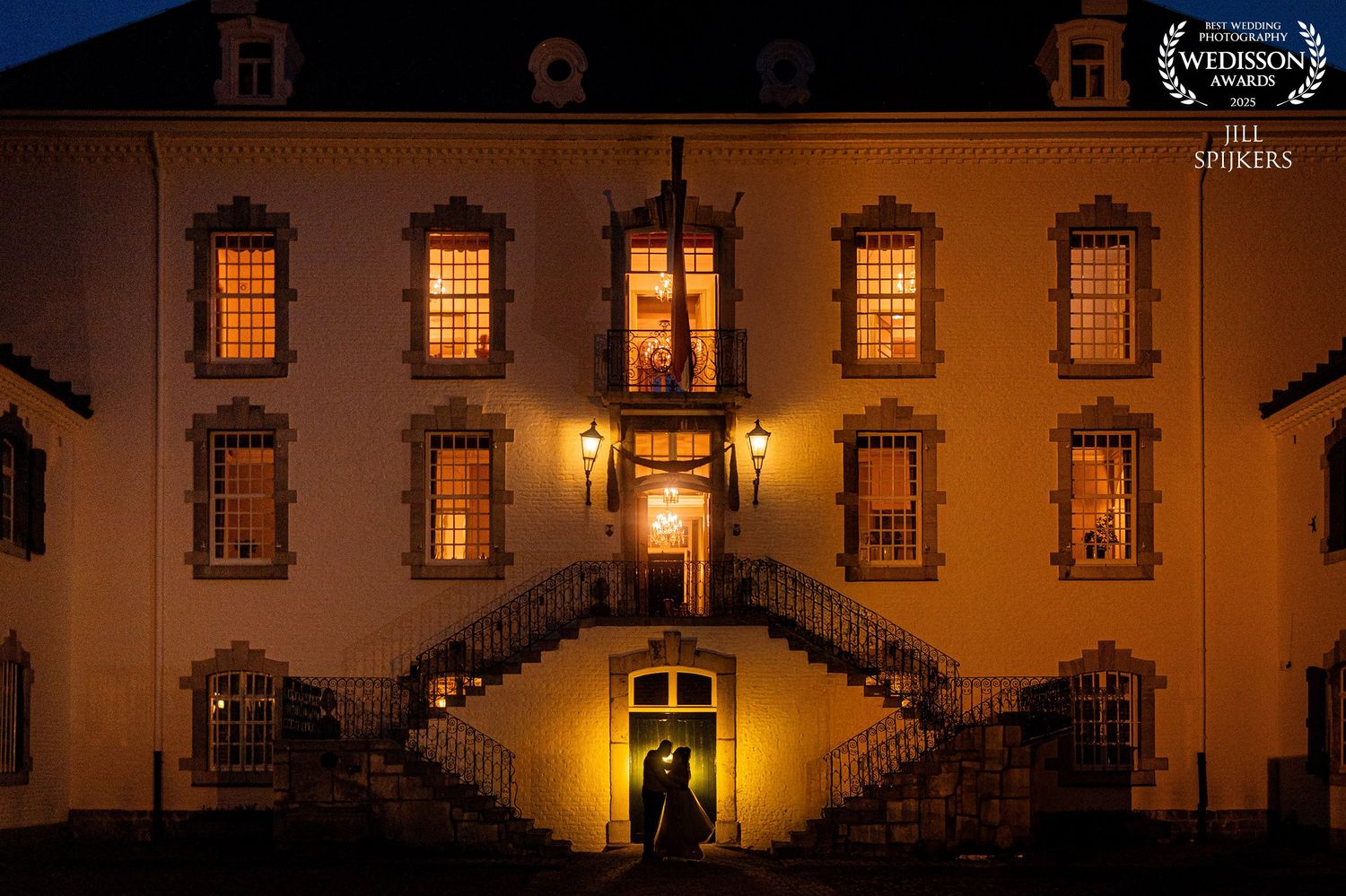 Historic mansion illuminated at night with glowing windows and curved double staircase entrance creating an elegant scene.