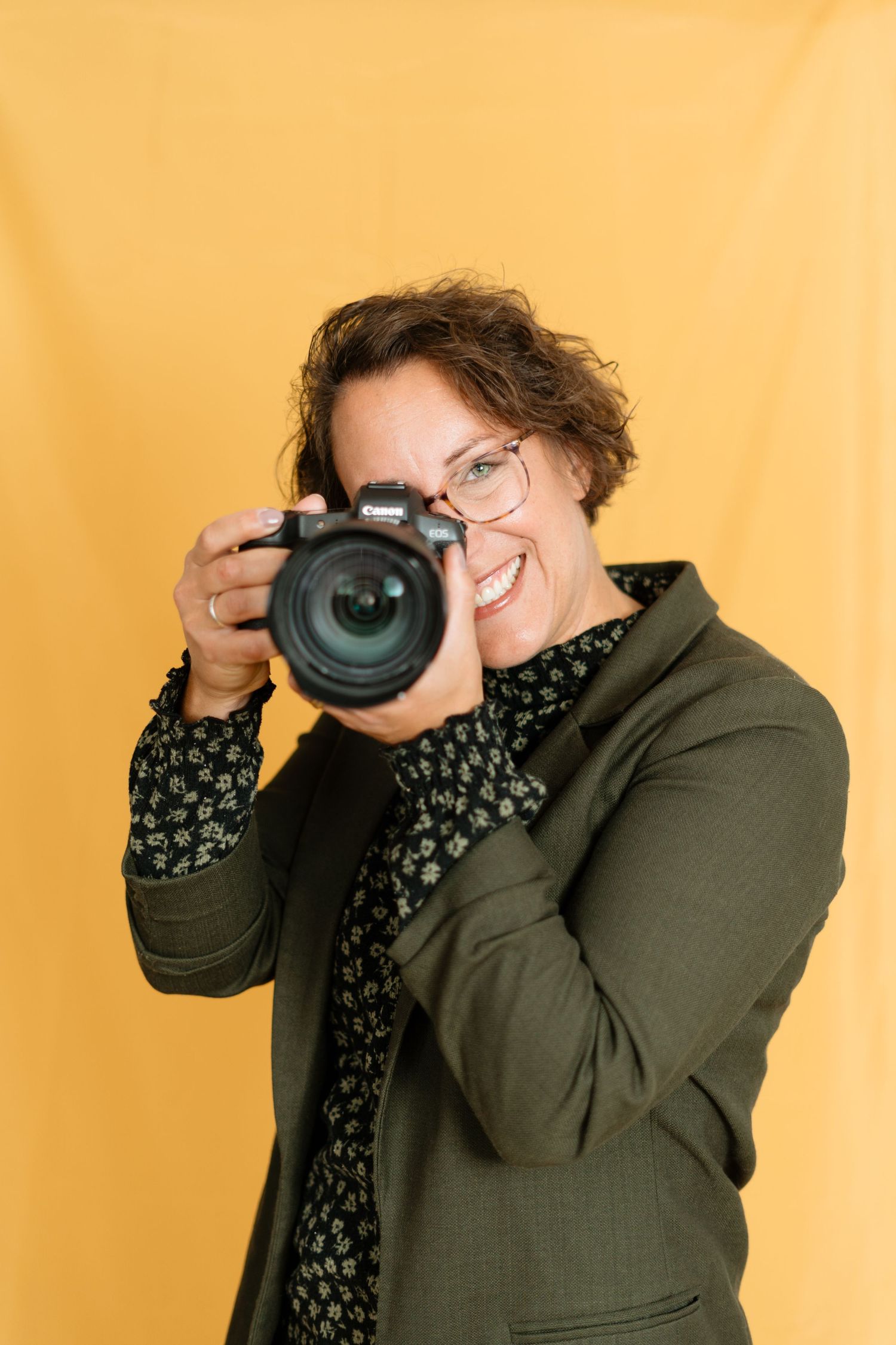 Professional photographer wearing glasses and olive green blazer holds DSLR camera against yellow background.