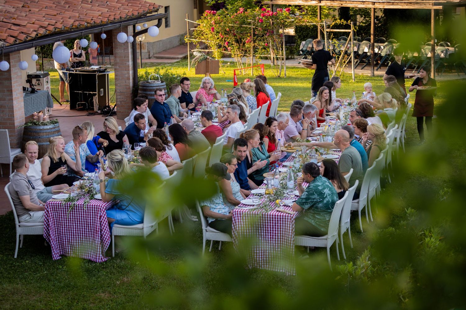 Guests dine at long outdoor tables during an elegant garden party with checkered tablecloths and white chairs.