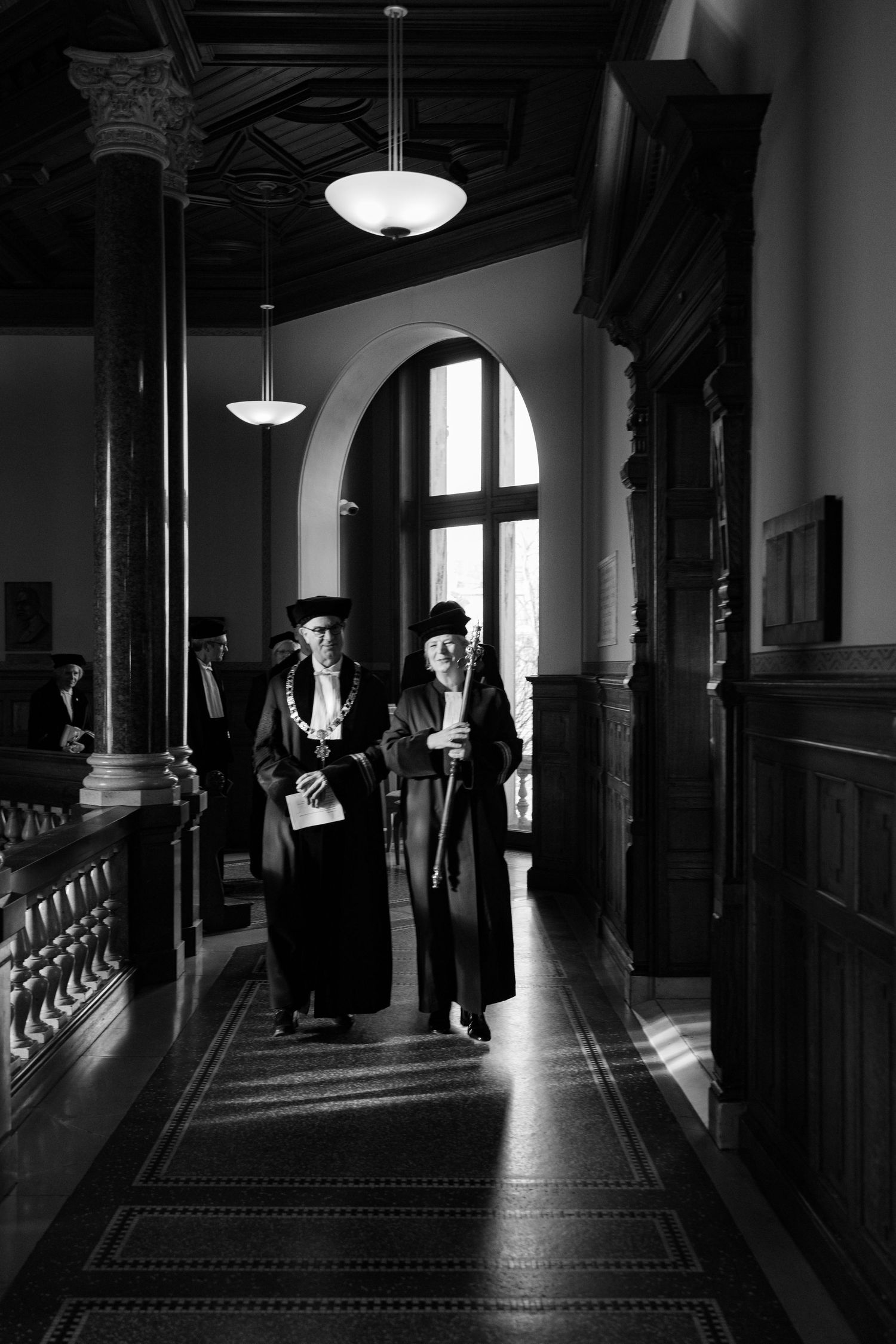 Two people in academic regalia walk down an ornate hallway with classical architecture.