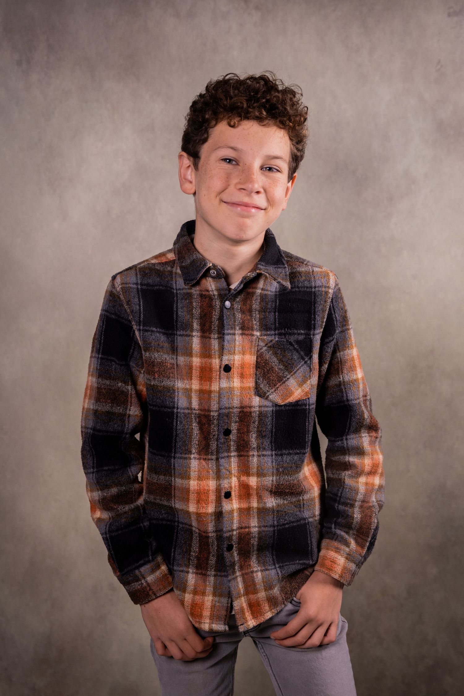 Child wearing plaid flannel shirt poses playfully against neutral studio background.