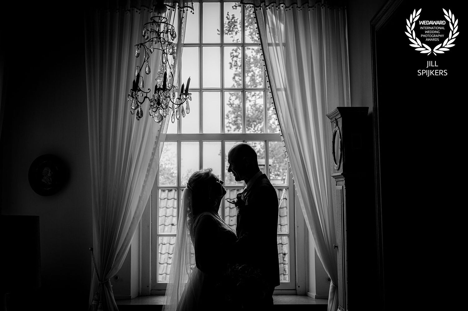 Silhouetted couple shares romantic moment by elegant window with chandelier.