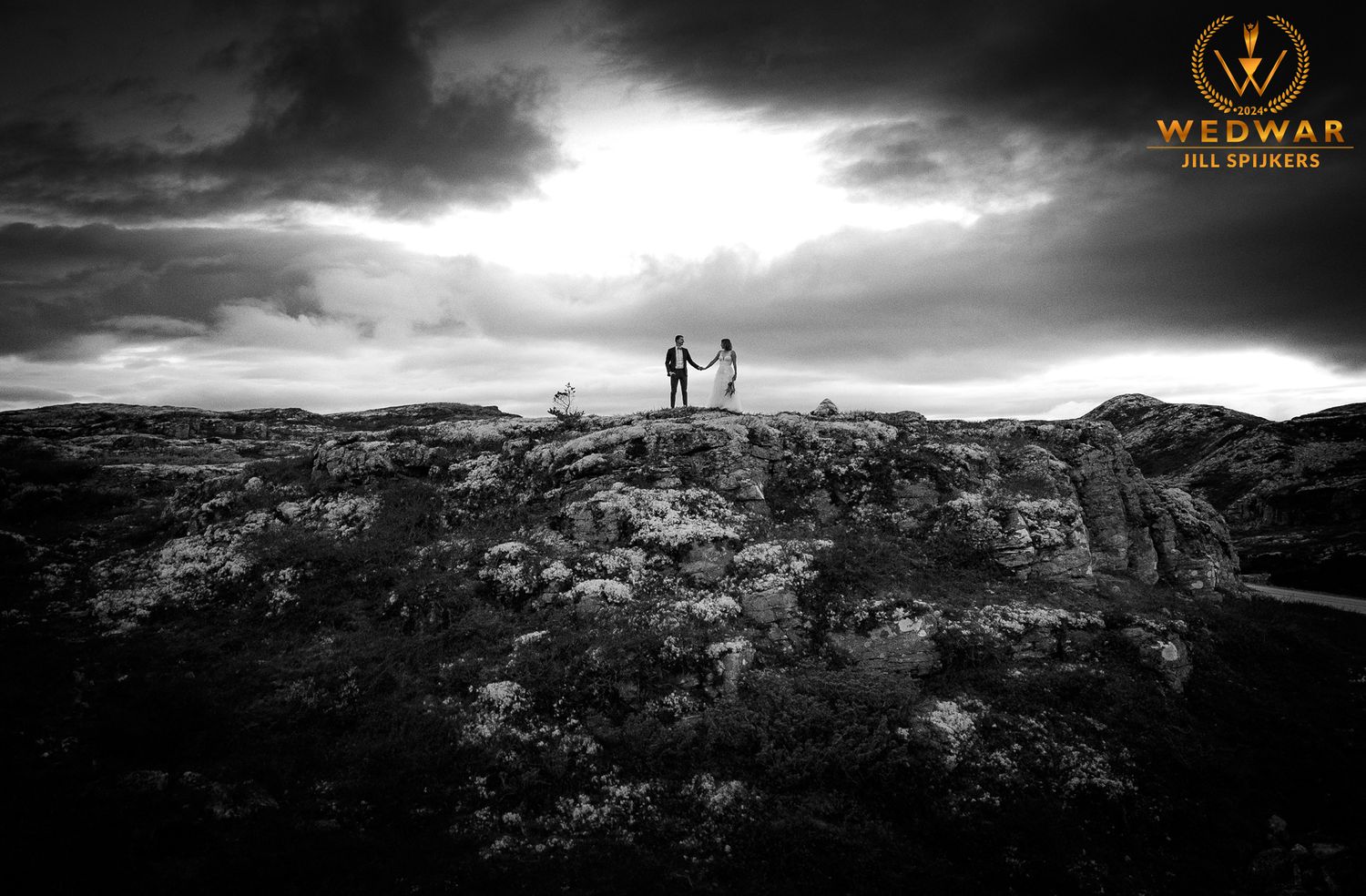 Silhouetted couple standing triumphantly on rocky mountaintop against moody sky.