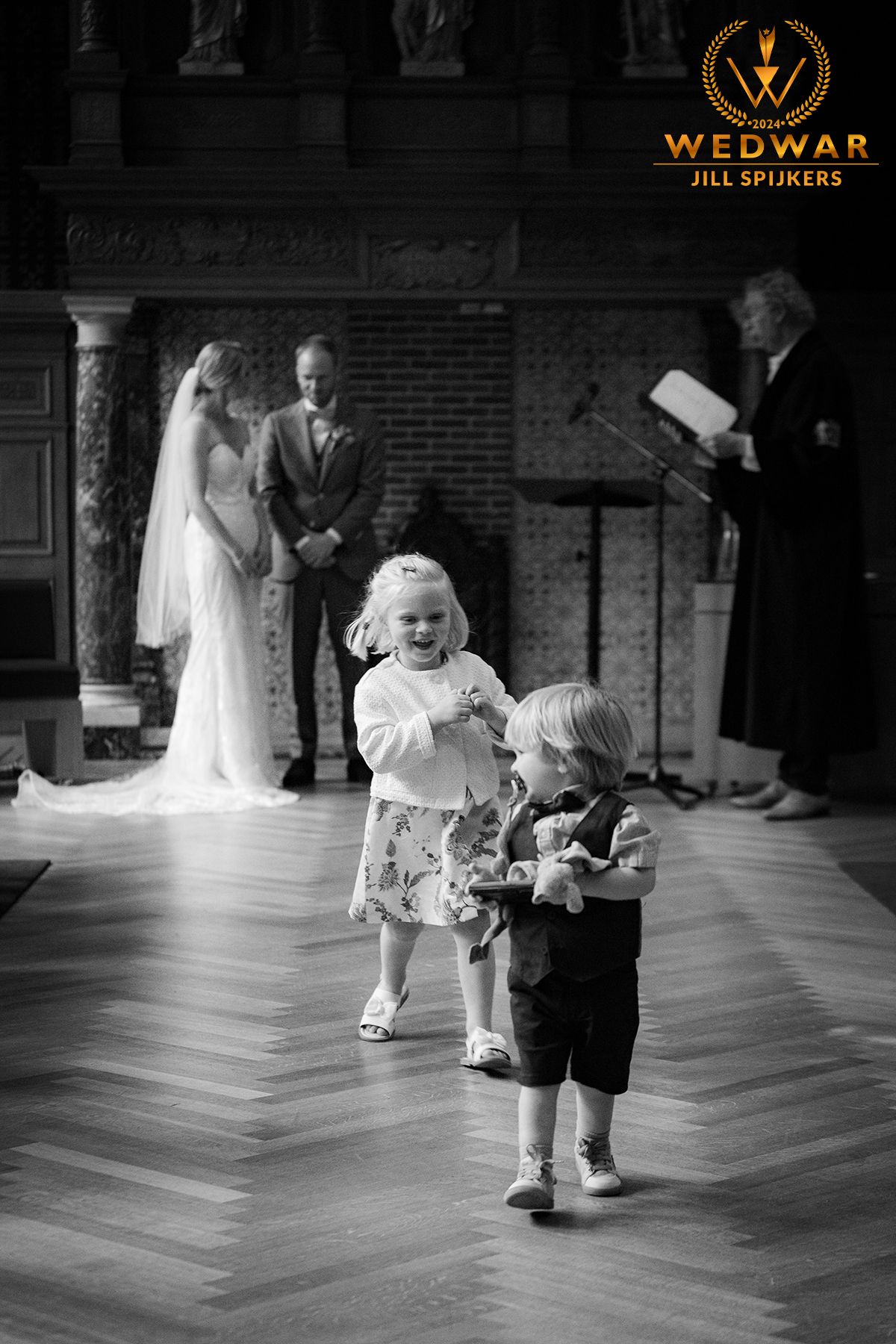 Black and white ceremony photo with toddlers in foreground and ceremony behind.
