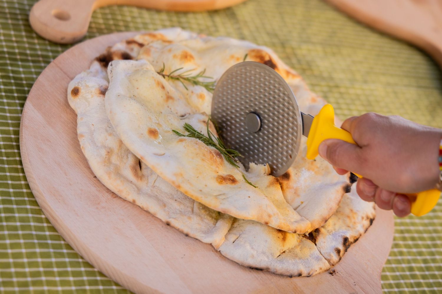 Someone using a pizza cutter to slice freshly baked flatbread garnished with herbs on a wooden board.