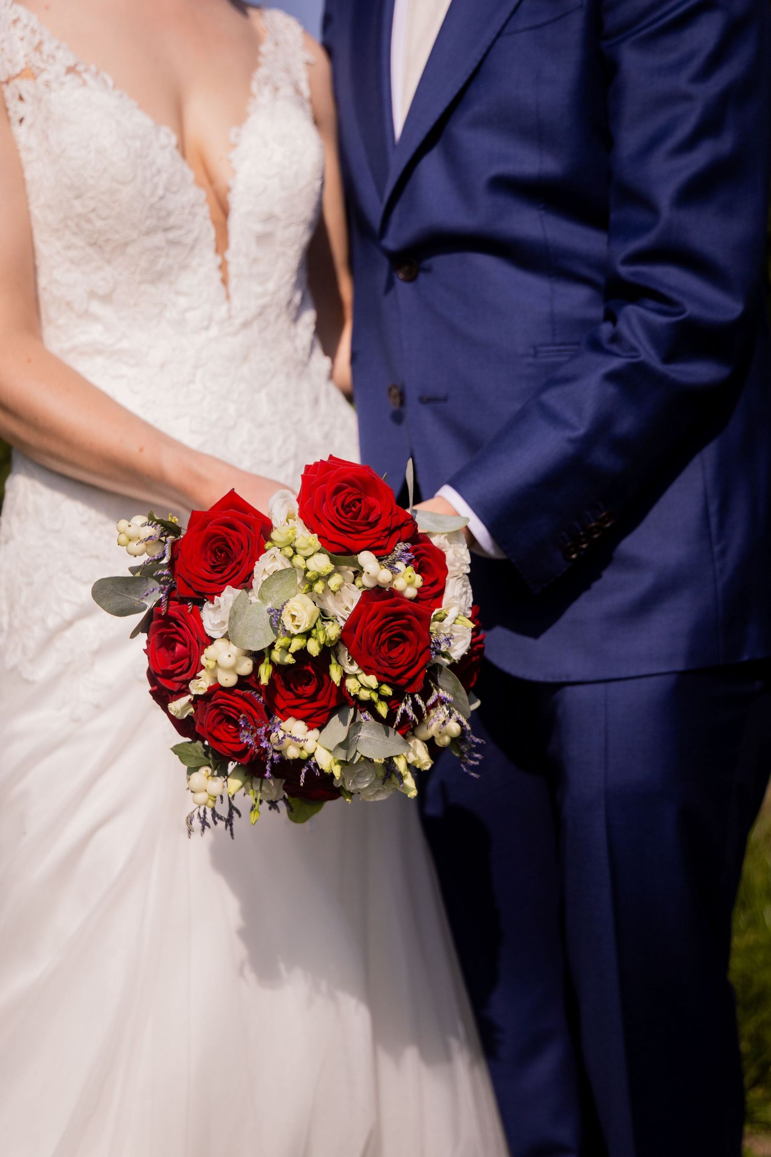 Wedding bouquet with red roses held between bride in lace dress and groom in navy suit.