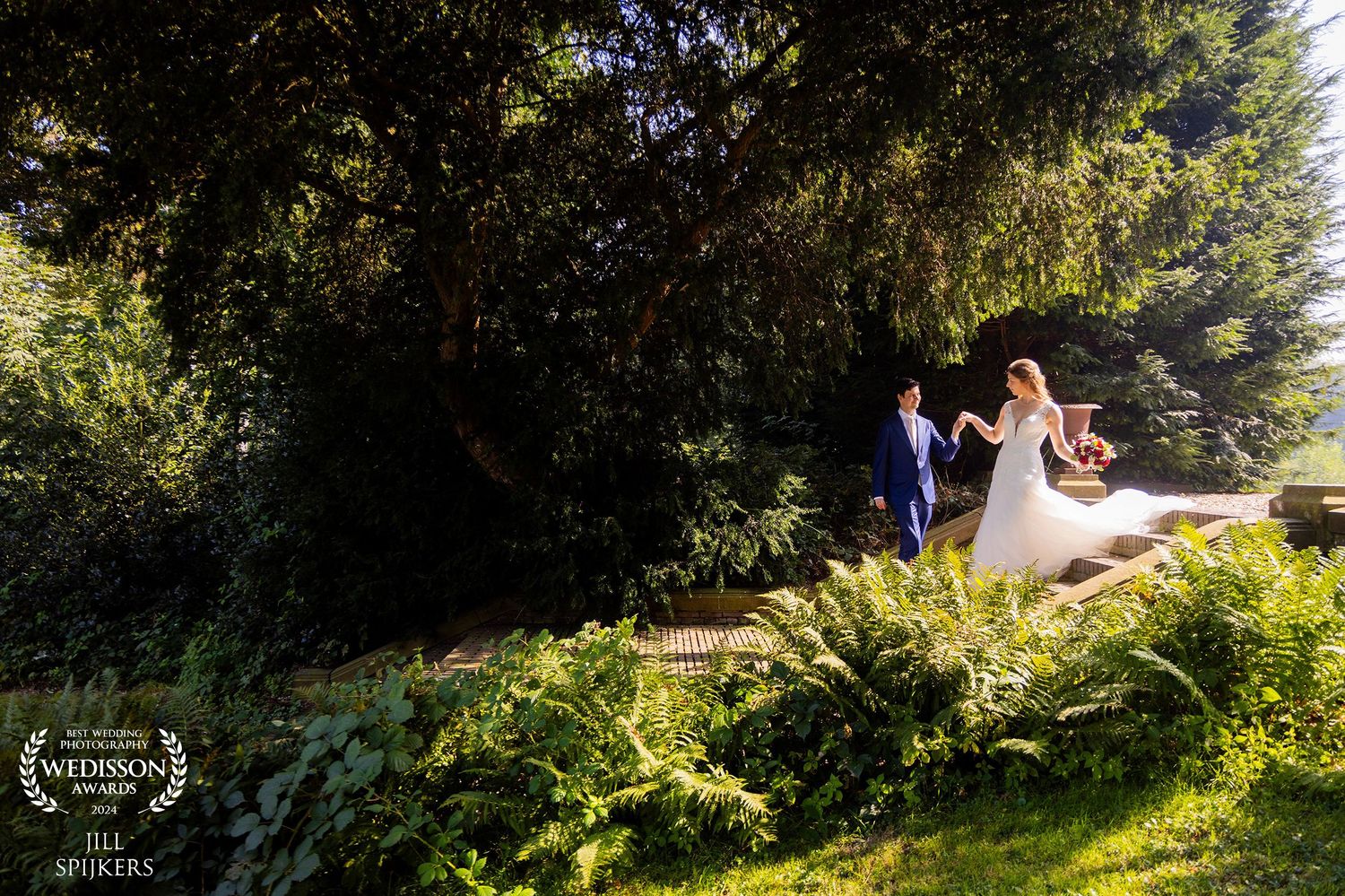 Wedding couple walks along garden path surrounded by lush greenery.