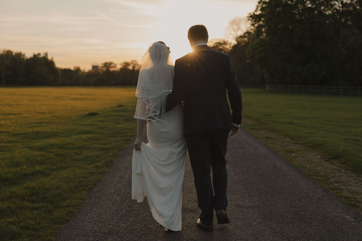 Silhouetted newlyweds walking down a path at sunset in a countryside setting.