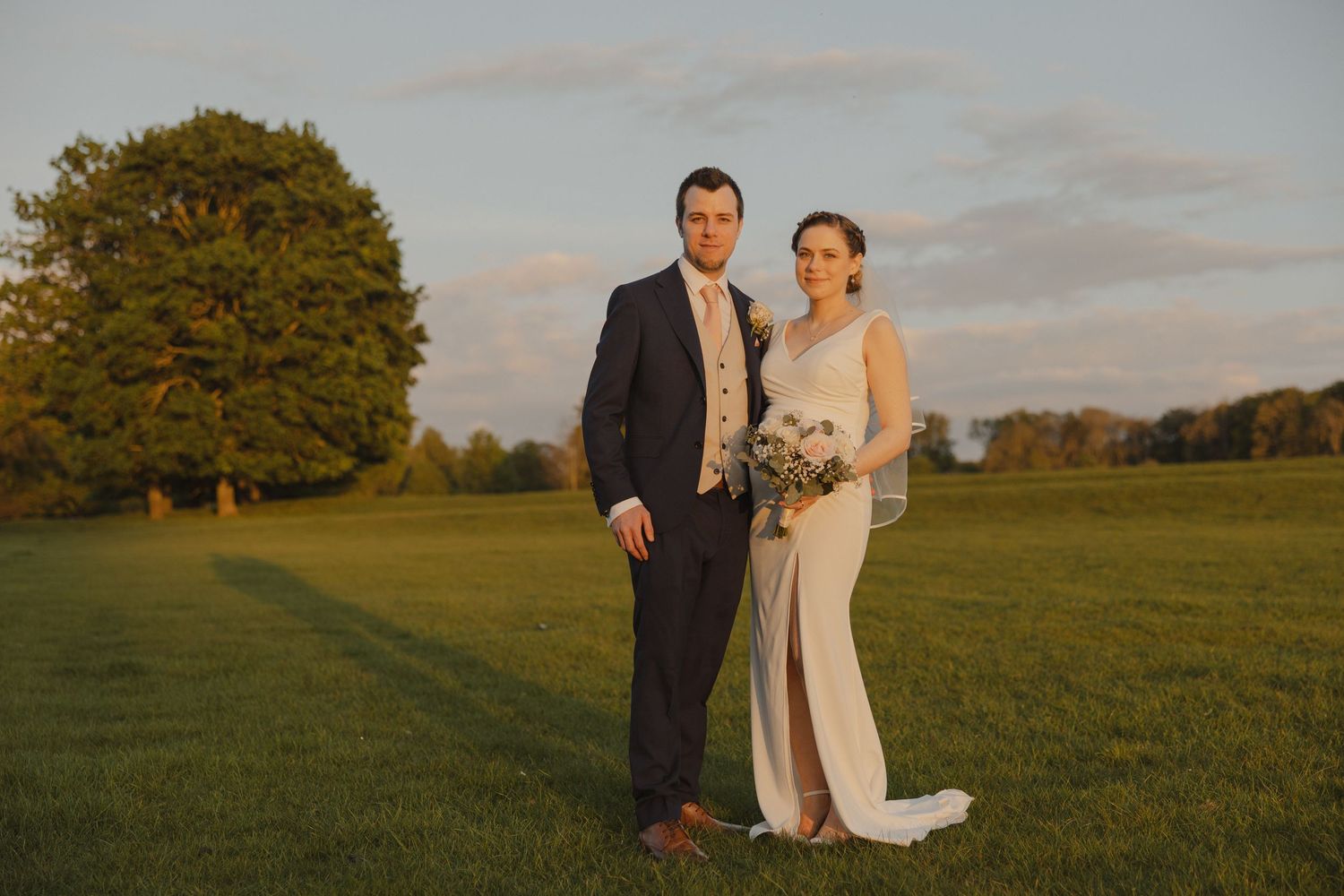 A wedding couple poses in formal attire on a grassy field during golden hour.
