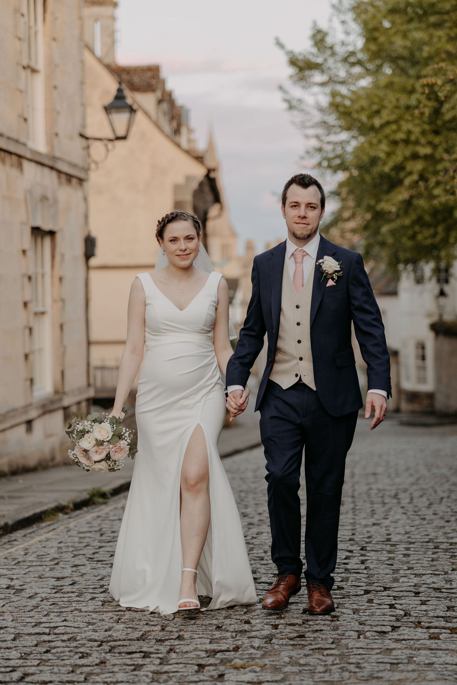 A wedding couple walks hand in hand down a cobblestone street wearing formal attire.