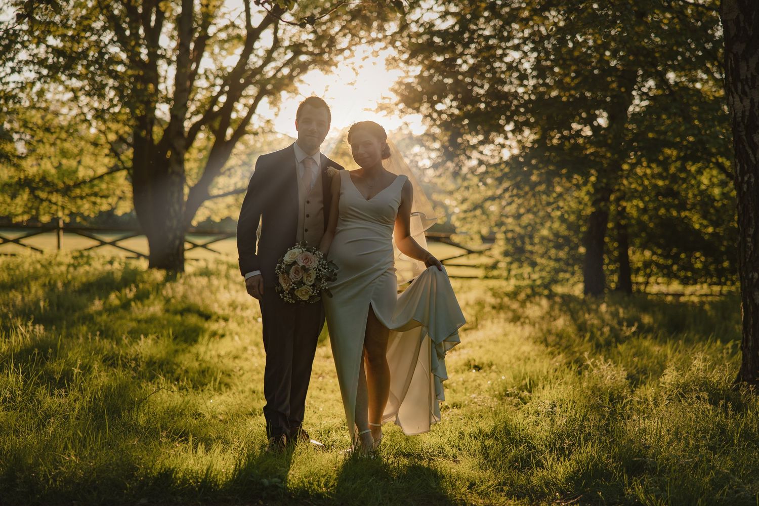 A couple walking hand in hand through a sun-dappled garden in wedding attire.