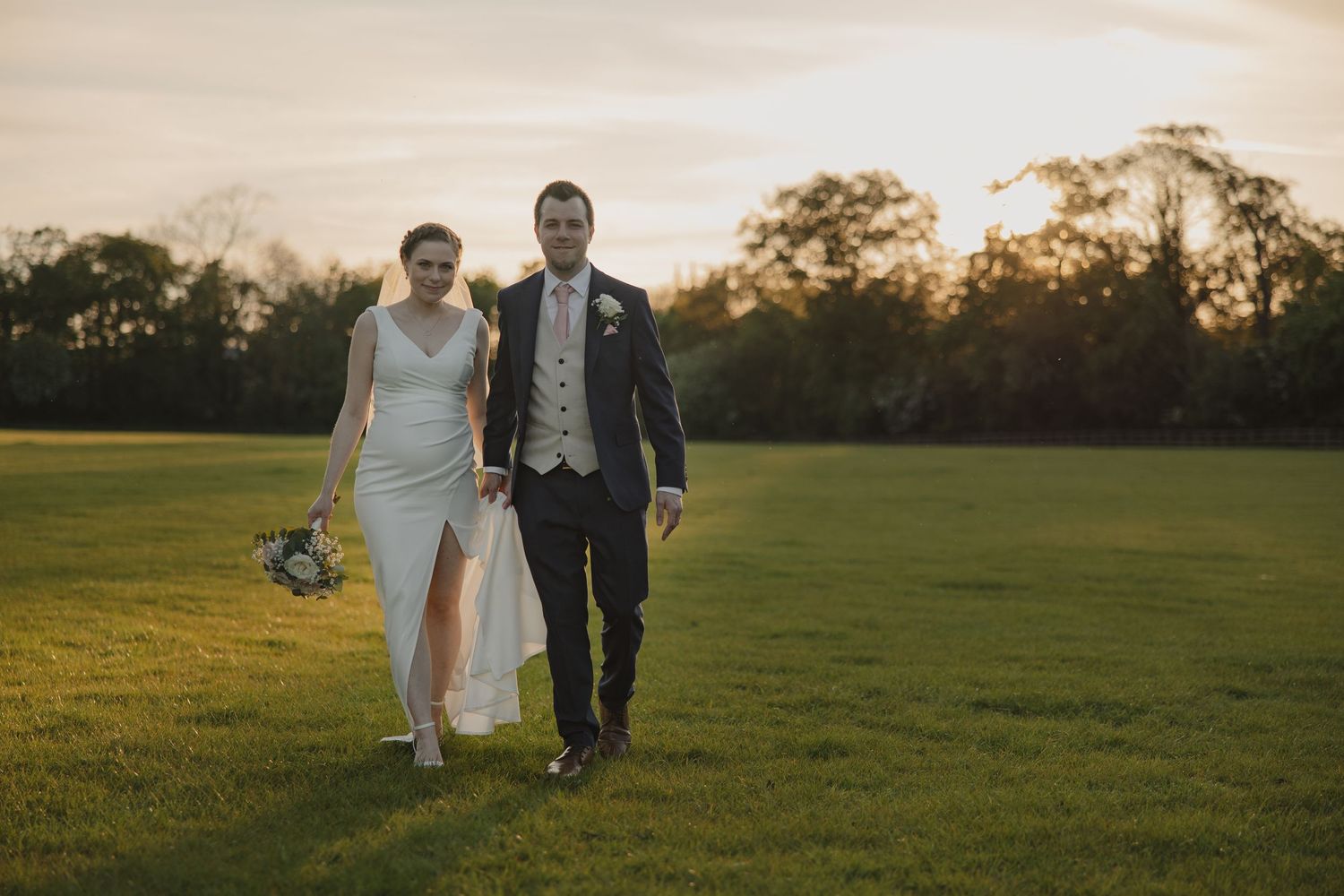 A couple walks hand in hand across a sunlit lawn at golden hour after their wedding ceremony.