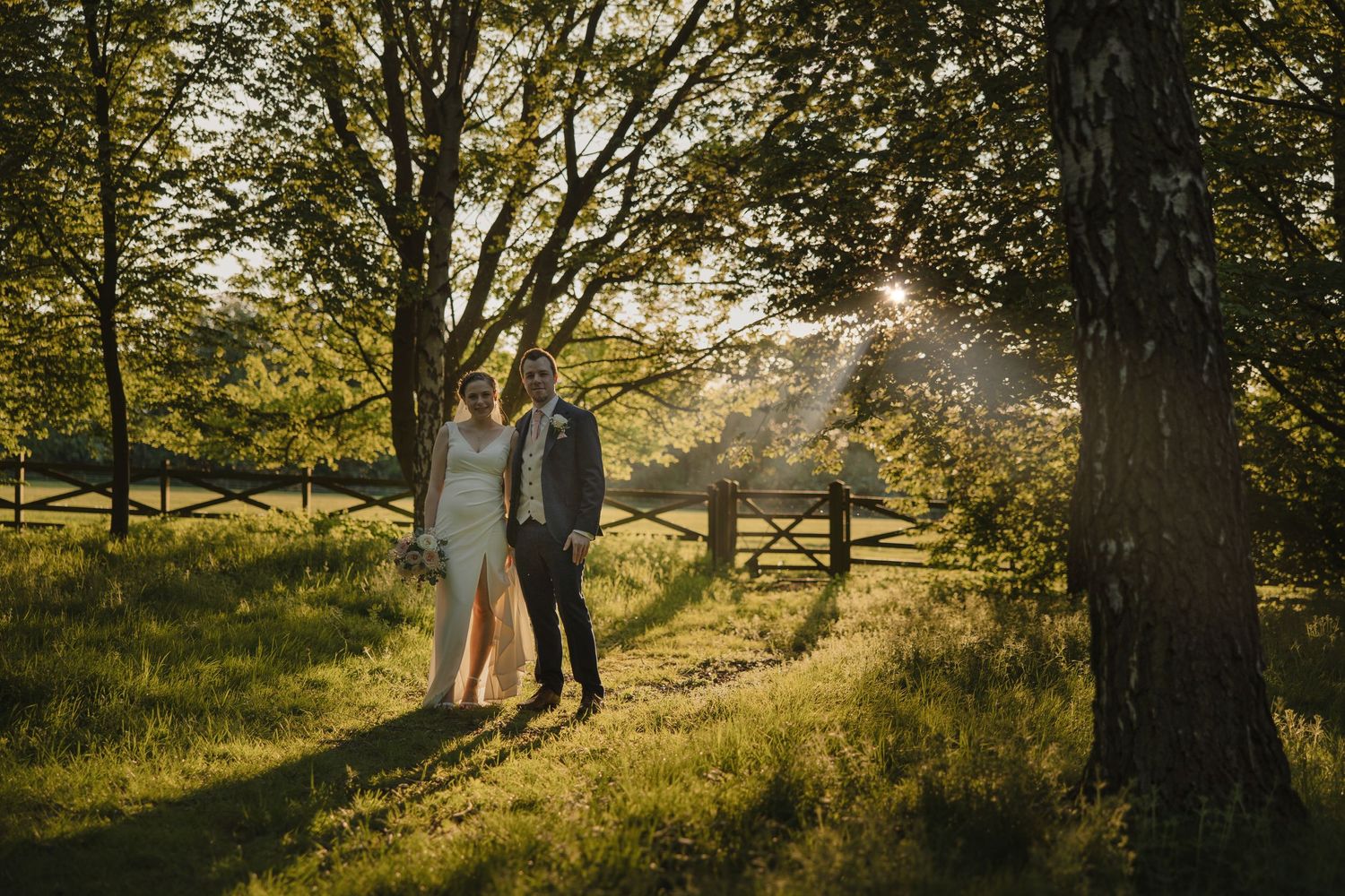 Newlyweds standing together on a sunlit path between trees with a wooden gate in the background.