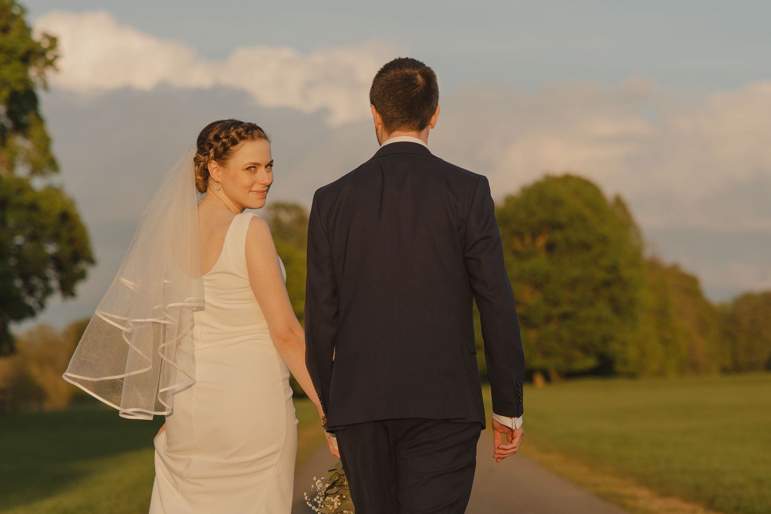 A newlywed couple walks through a scenic garden during sunset with a flowing veil trailing behind.