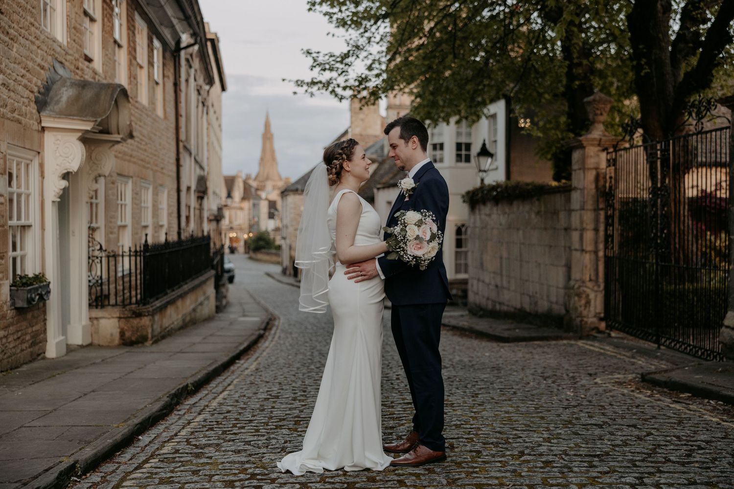 Wedding couple stands on cobblestone street with historic buildings and church spire backdrop.