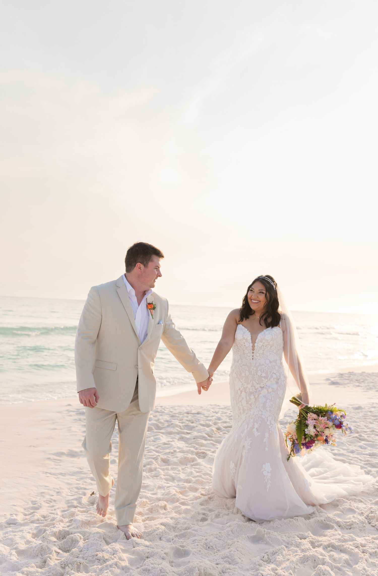 A couple walks hand in hand on a white sand beach at sunset, wearing wedding attire and a bridal bouquet.