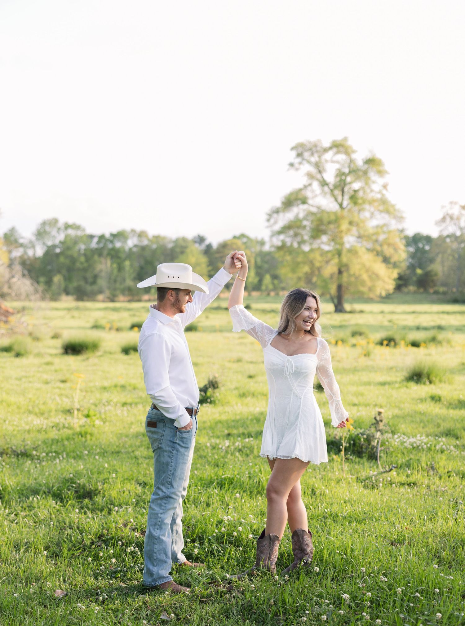 A couple in western attire dance together in a sunlit meadow surrounded by green grass and trees at golden hour.