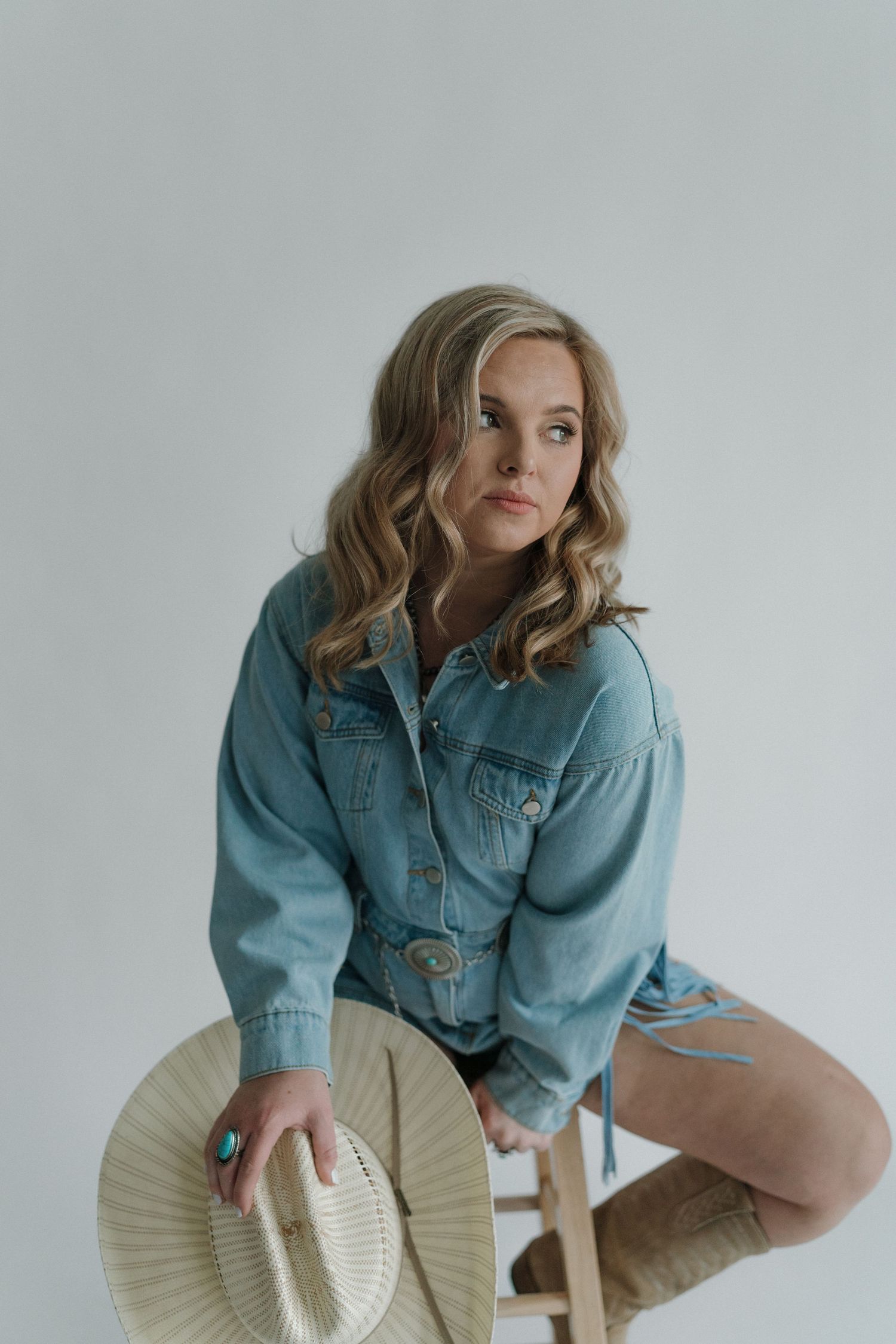Stylish denim jacket model sits on stool holding straw hat against minimalist grey background in studio portrait.