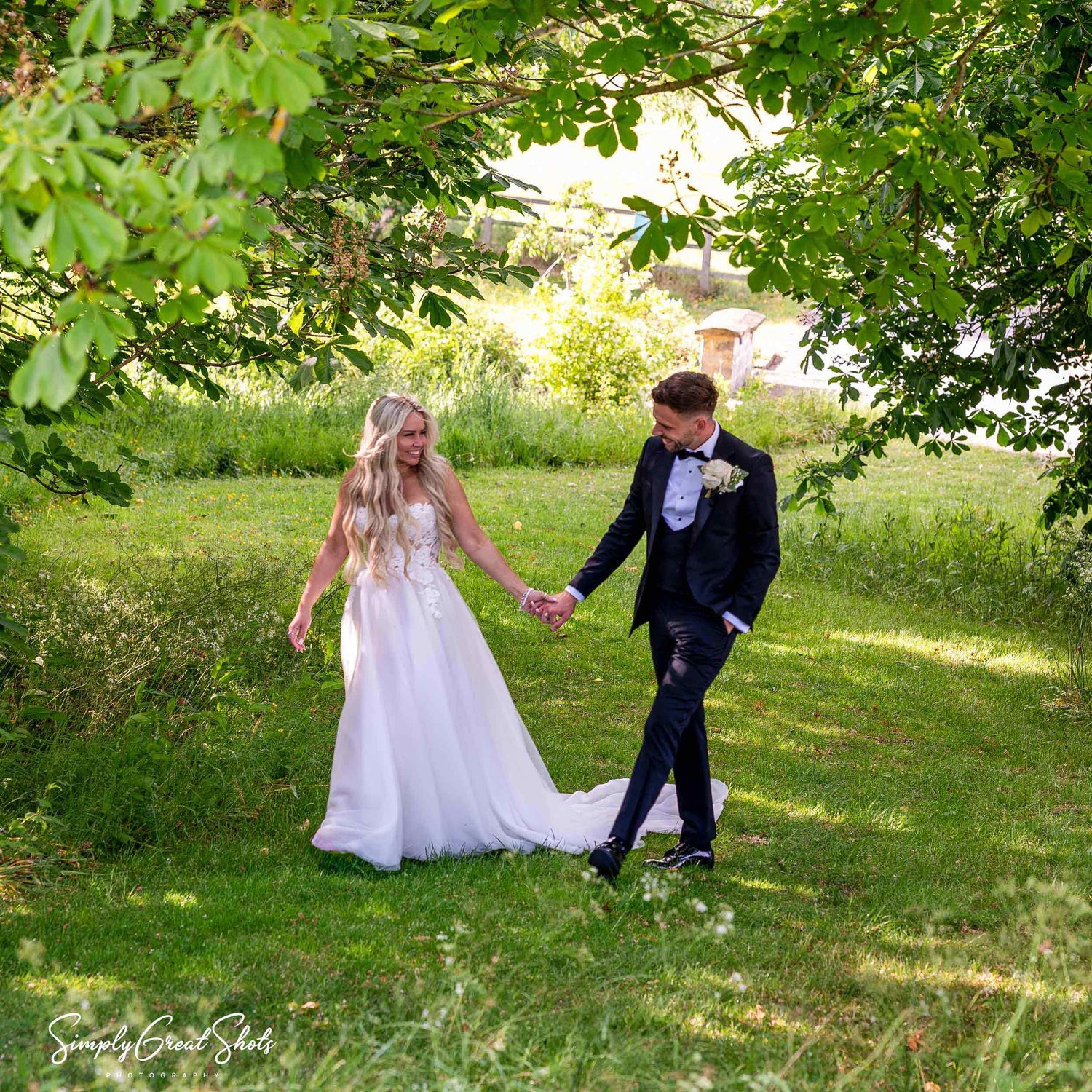 A couple in wedding attire holds hands while walking through a lush garden at Blackwell Grange on a sunny afternoon.