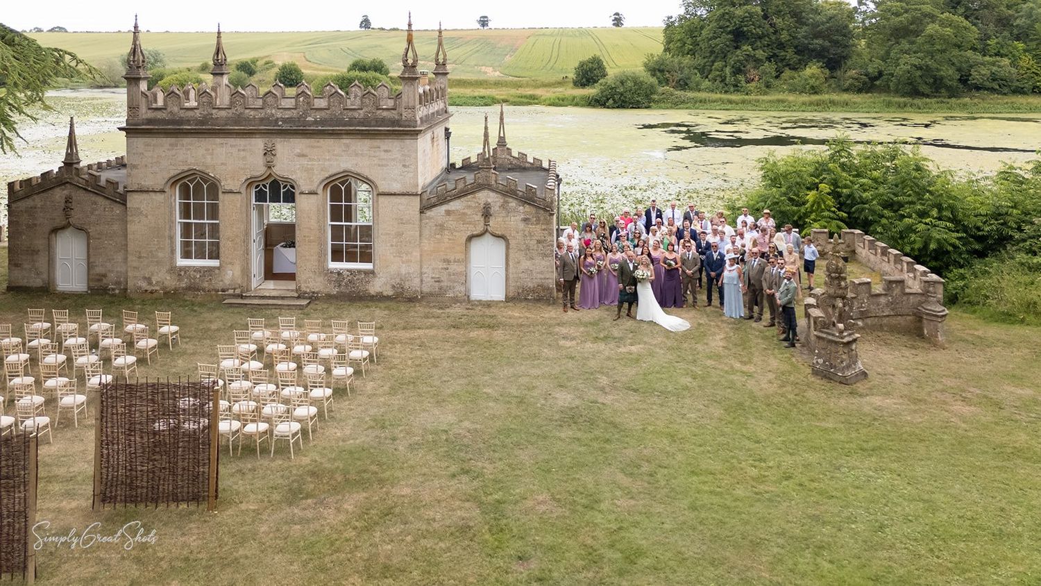 A wedding party gathers on the lawn to the side of a small Gothic-style stone building with chairs set up for an outdoor ceremony.