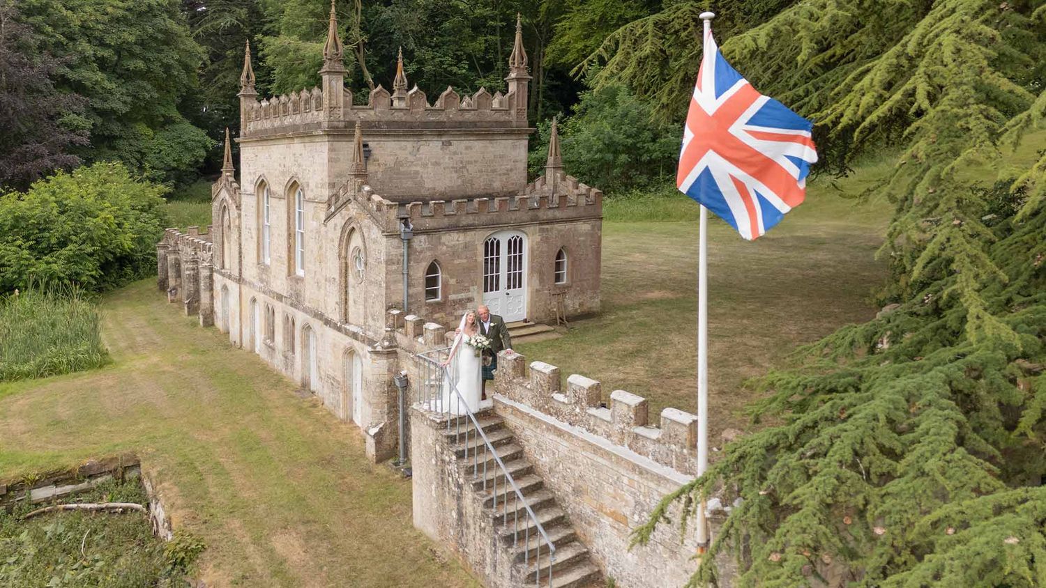 Fort Henry, a stone country manor house with a British flag flying, surrounded by green gardens viewed from an elevated angle.