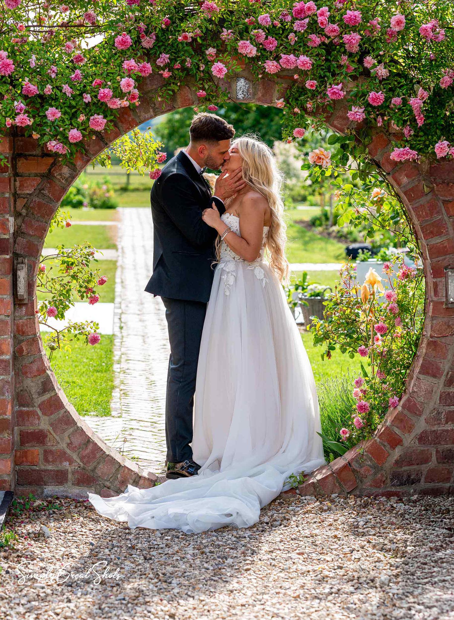 A wedding couple stand beneath a circular brick archway adorned with blooming pink climbing roses in a garden setting at Blackwell Grange.