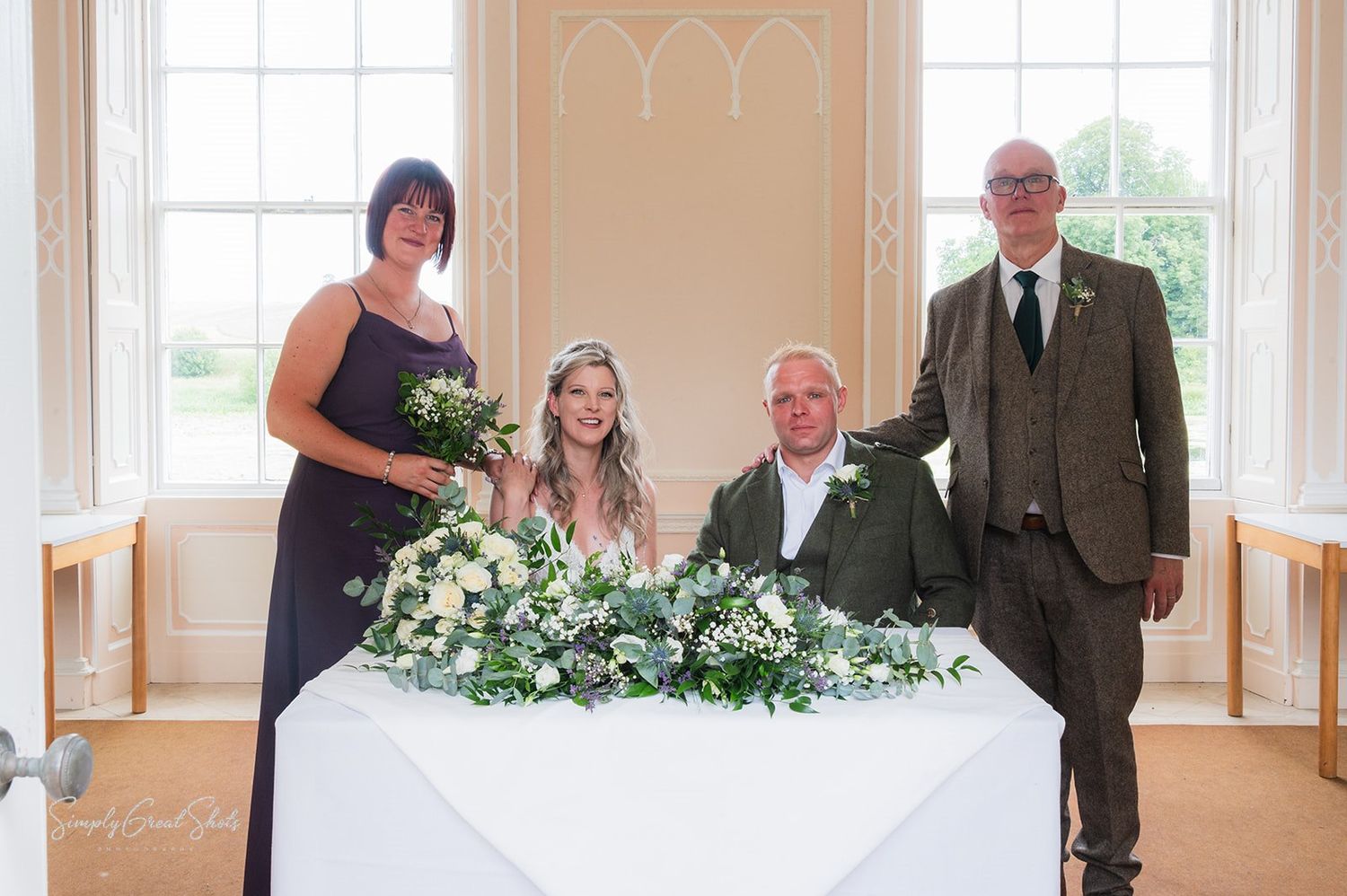 Wedding ceremony party stands at decorated registry table with floral arrangement in elegant room with arched windows.
