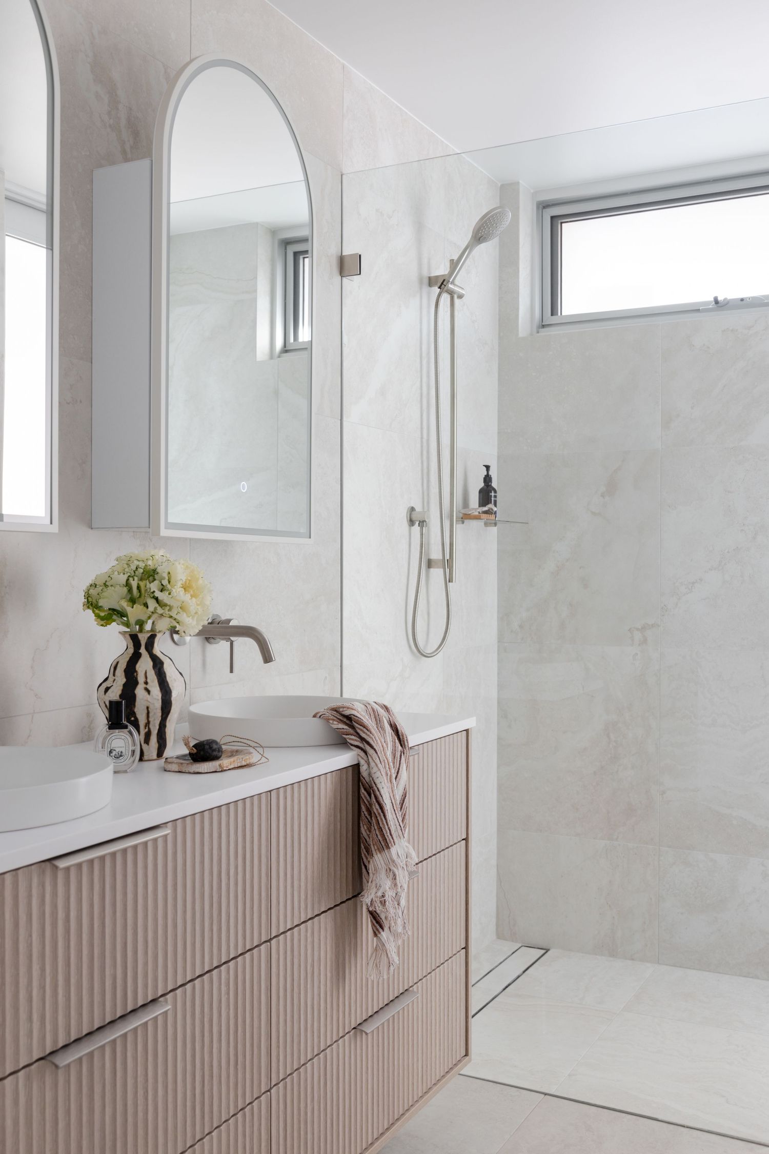 Modern white bathroom featuring a taupe ribbed vanity, oval mirror, and sleek walk-in shower with high windows.