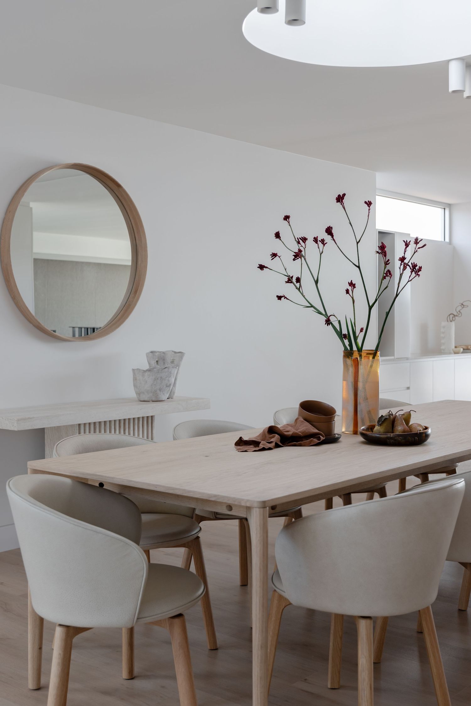 Minimalist dining area with round mirror, wooden table and white upholstered chairs.