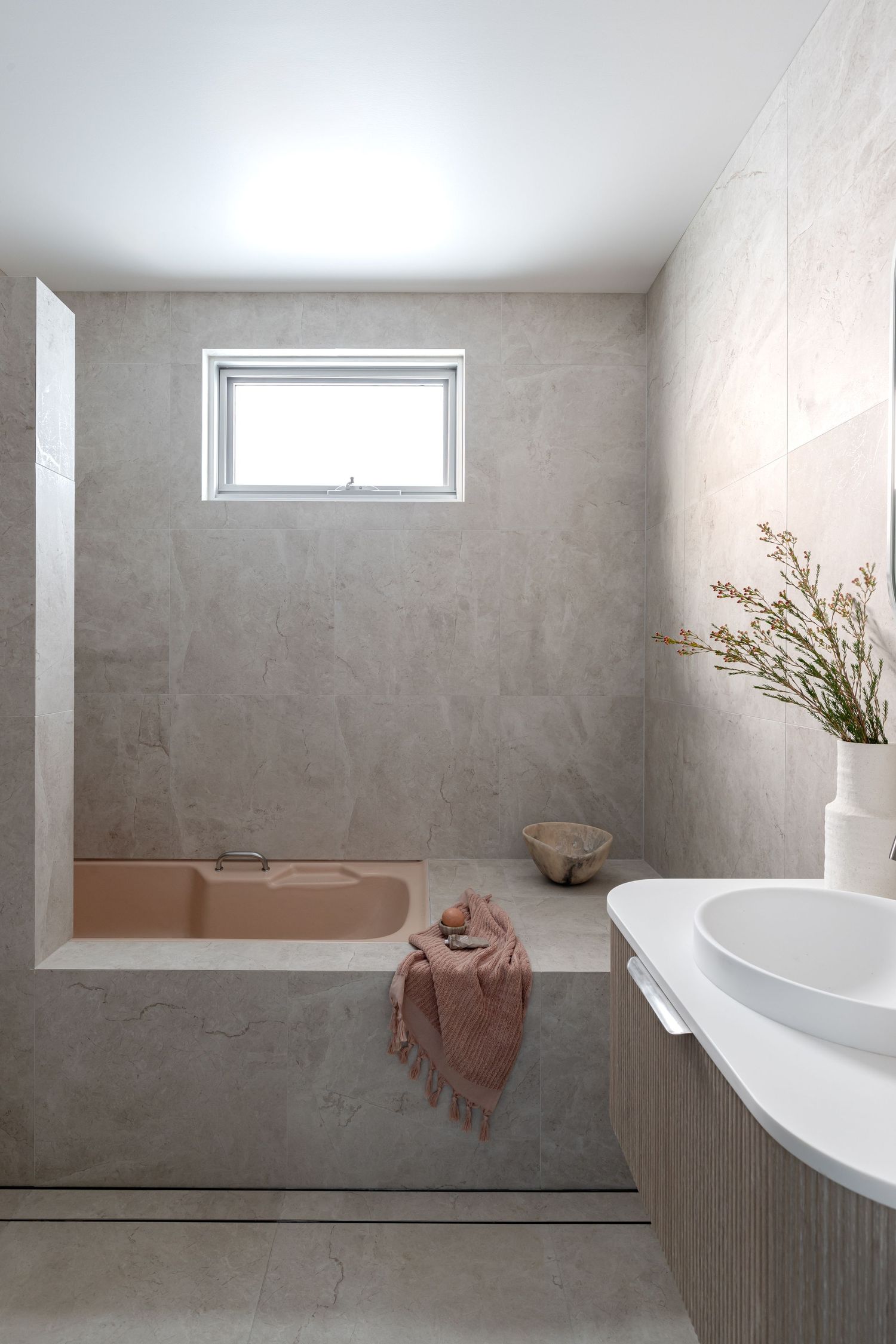 Modern bathroom with pink bathtub, high window, and minimalist grey tile design.