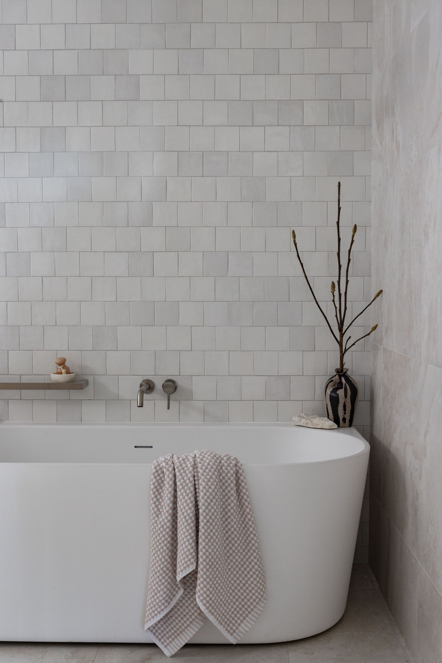 Freestanding white bathtub against subway tile wall with decorative branches in a vase.