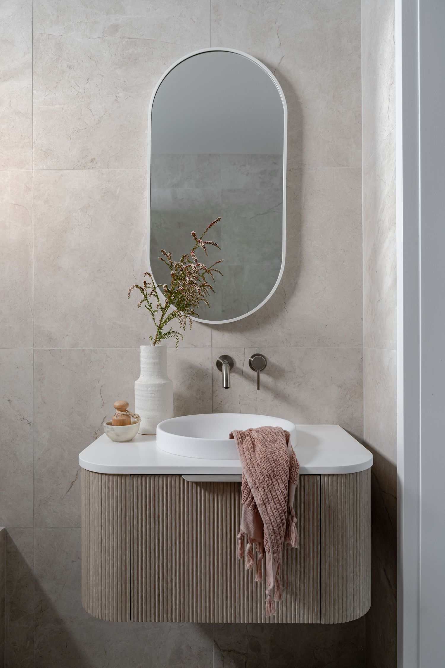 Sleek bathroom vanity with oval mirror, floating cabinet, and pink accents against grey walls.