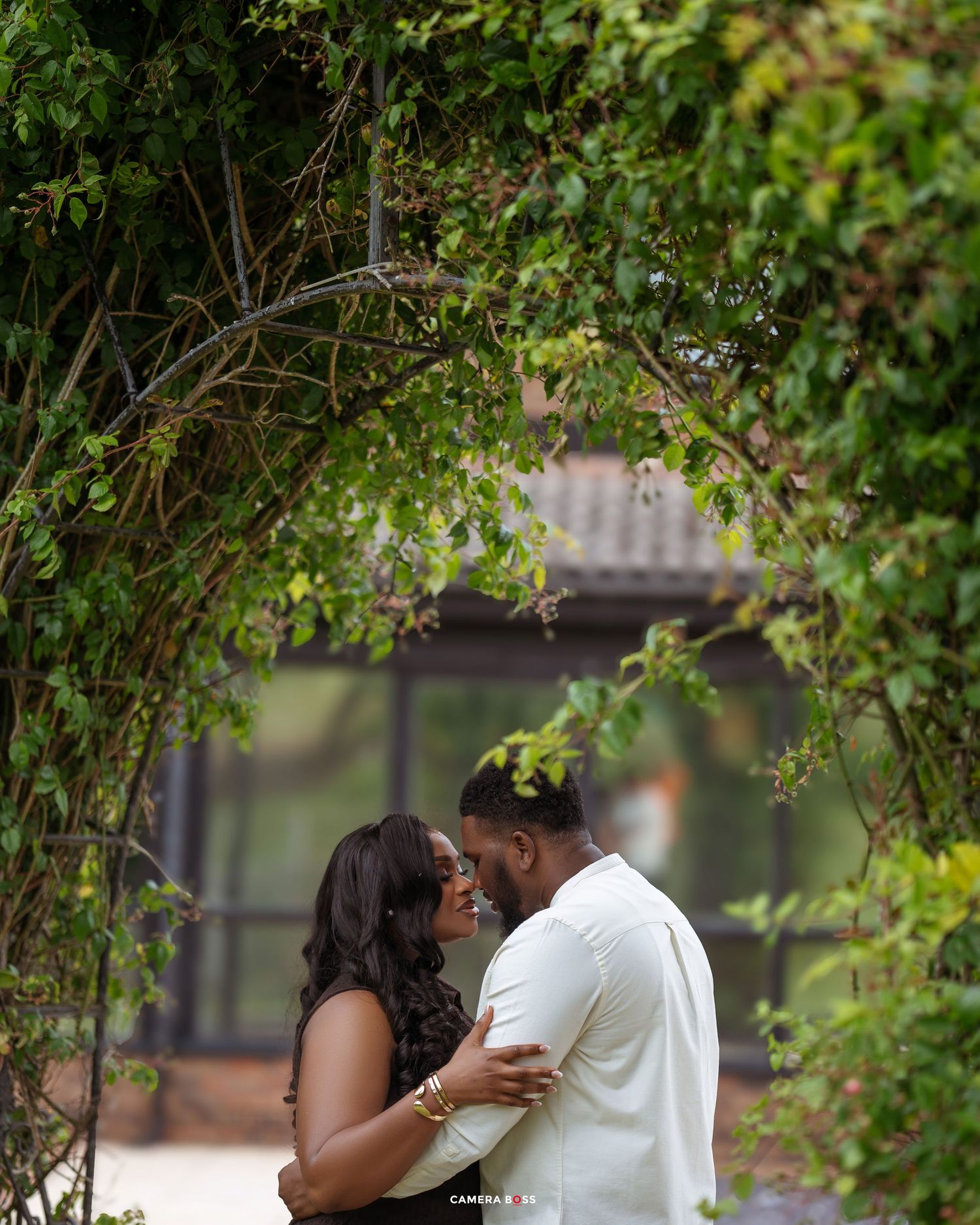 A couple share an intimate moment under a natural green archway in a garden setting.