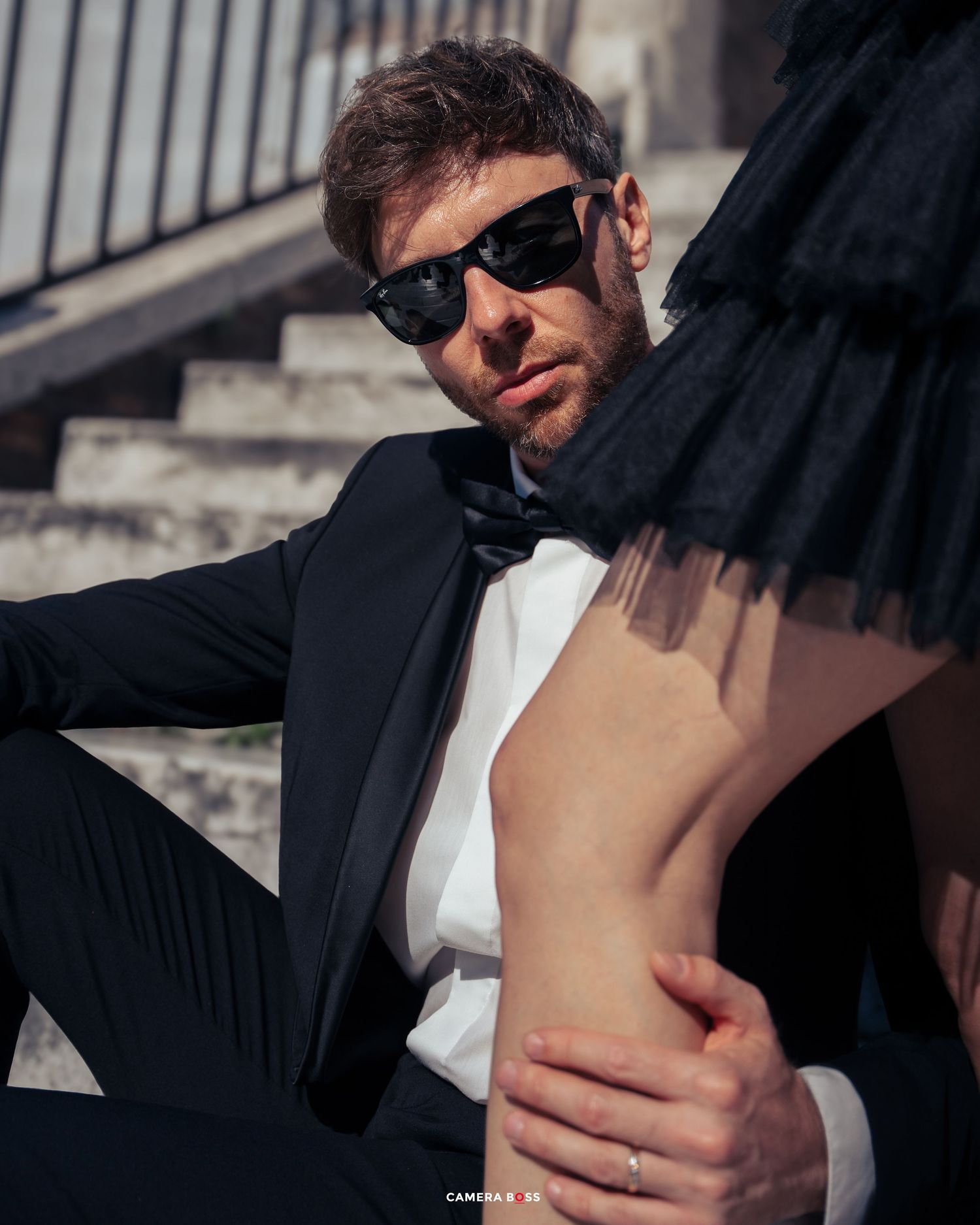 A person in a black tuxedo and sunglasses poses on stone steps during a stylish outdoor photo session.