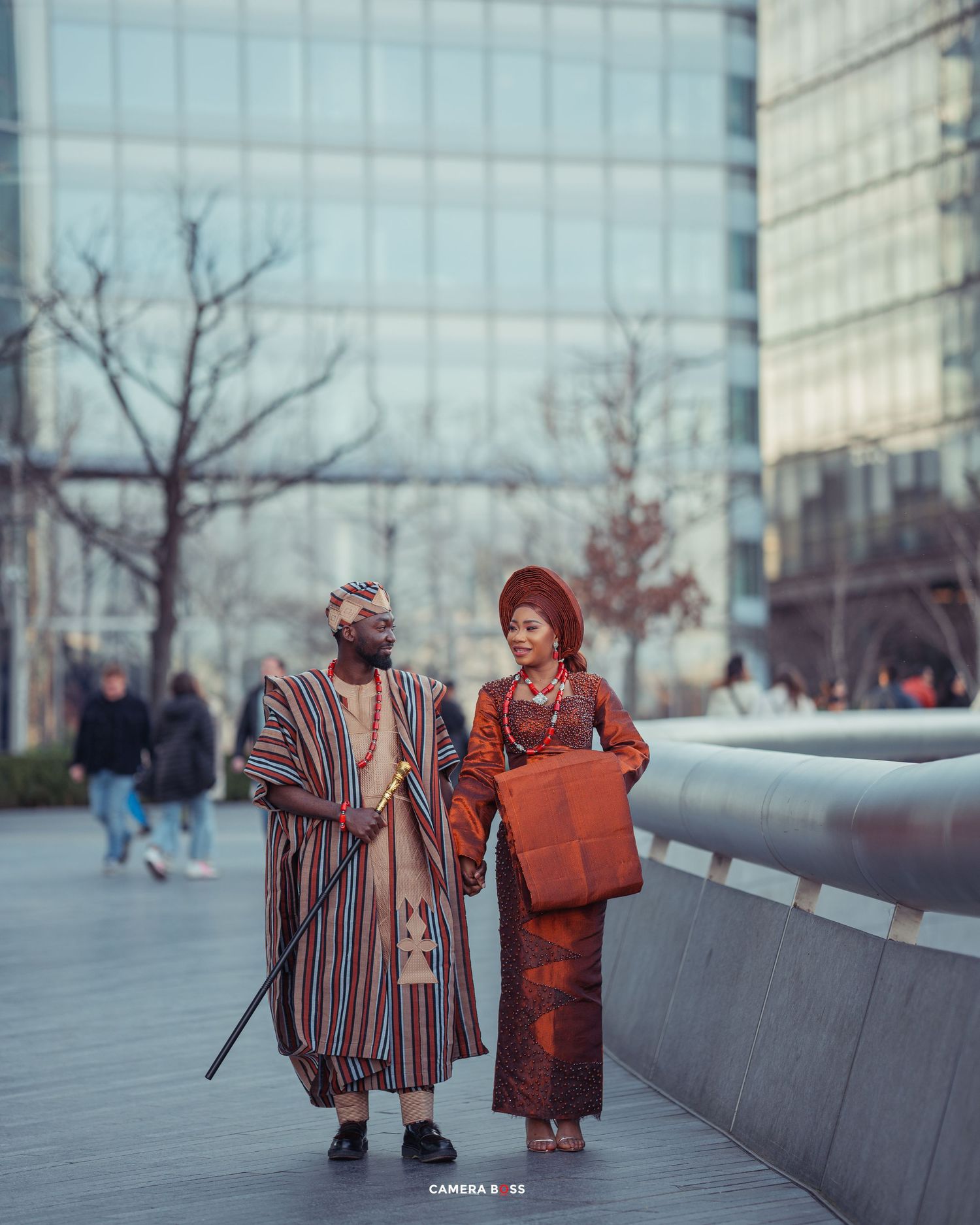 Two fashionably dressed figures in vibrant traditional outfits stand together on a modern city sidewalk near glass buildings.