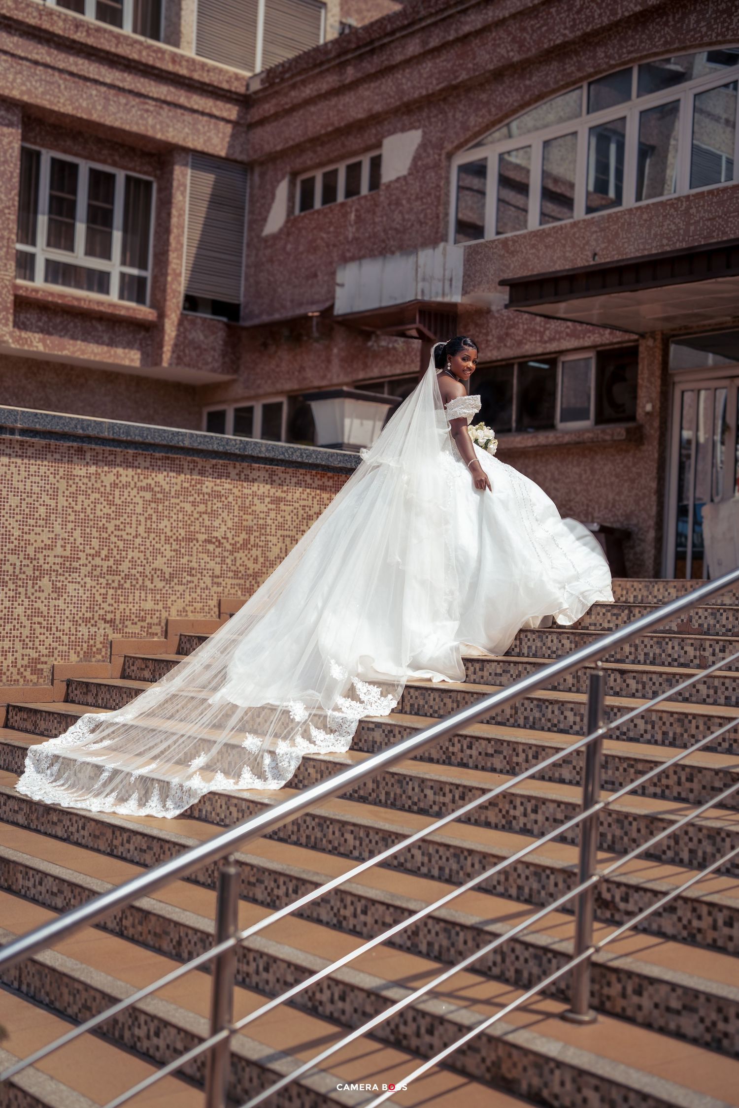 A wedding dress with a flowing train cascades down ornate stone steps in front of a brick building.