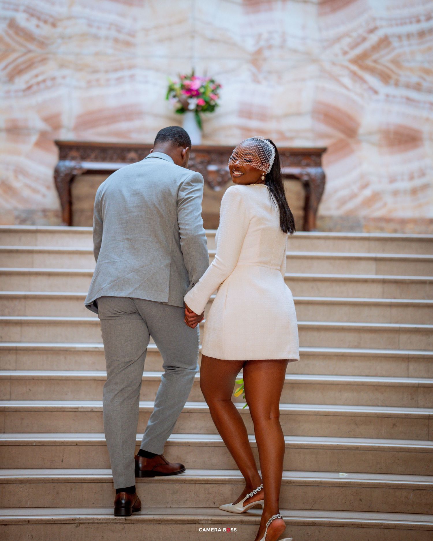Couple walking up elegant stone stairs with decorative walls and floral arrangement, dressed in gray suit and white outfit.
