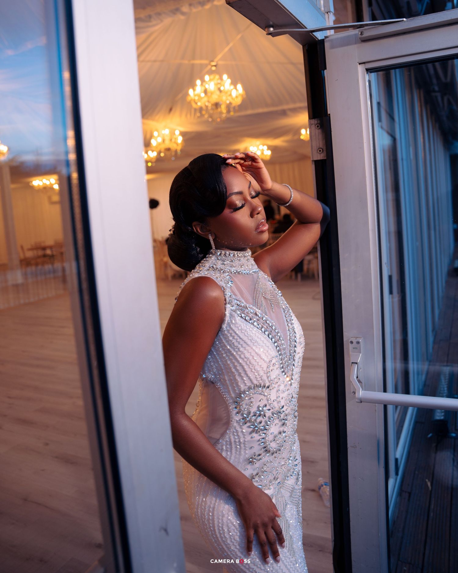 Elegant lace wedding dress silhouetted in doorway with crystal chandeliers glowing in luxurious ballroom background.