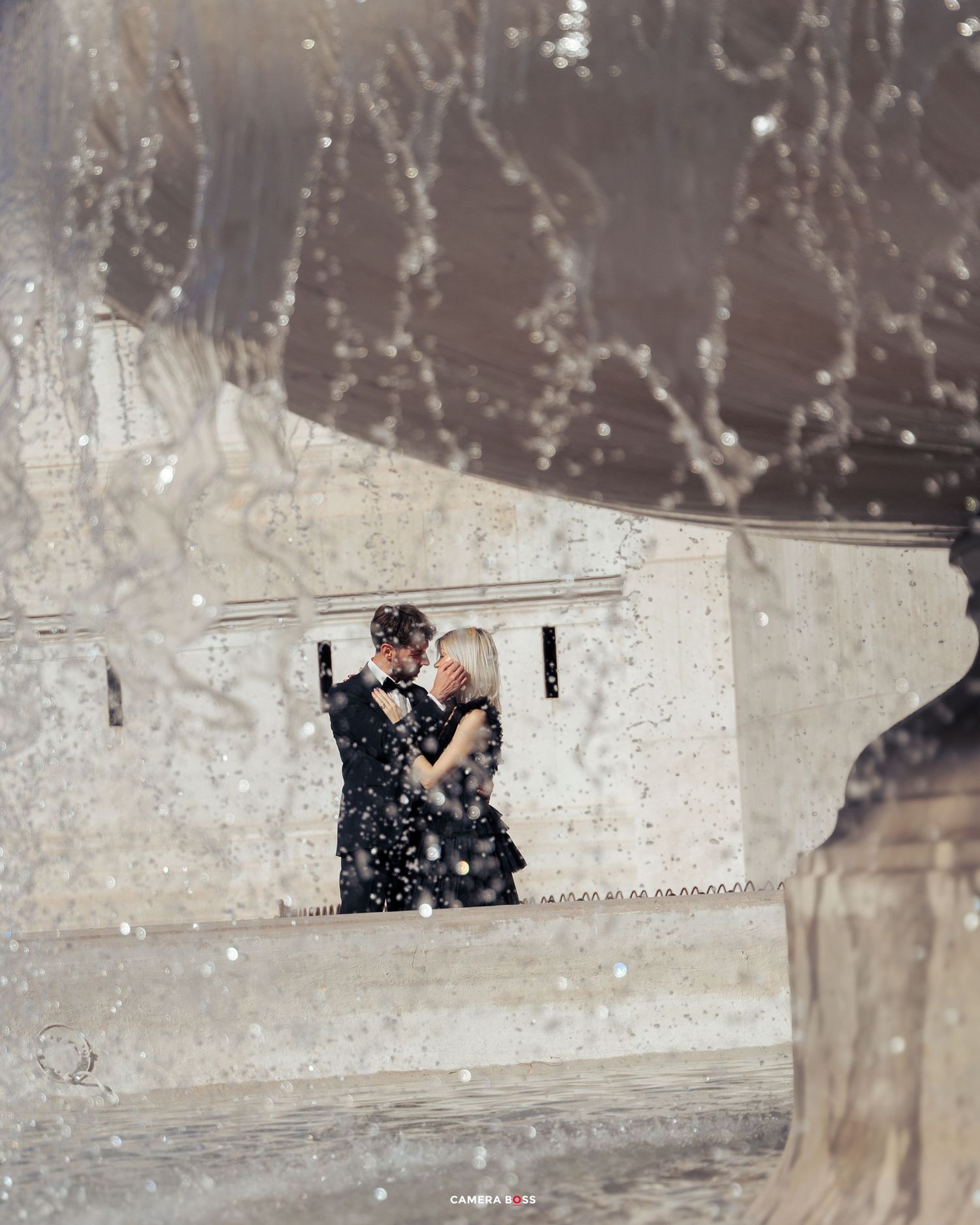 Couple embraces in window frame while soft white snow falls around them in romantic winter scene.