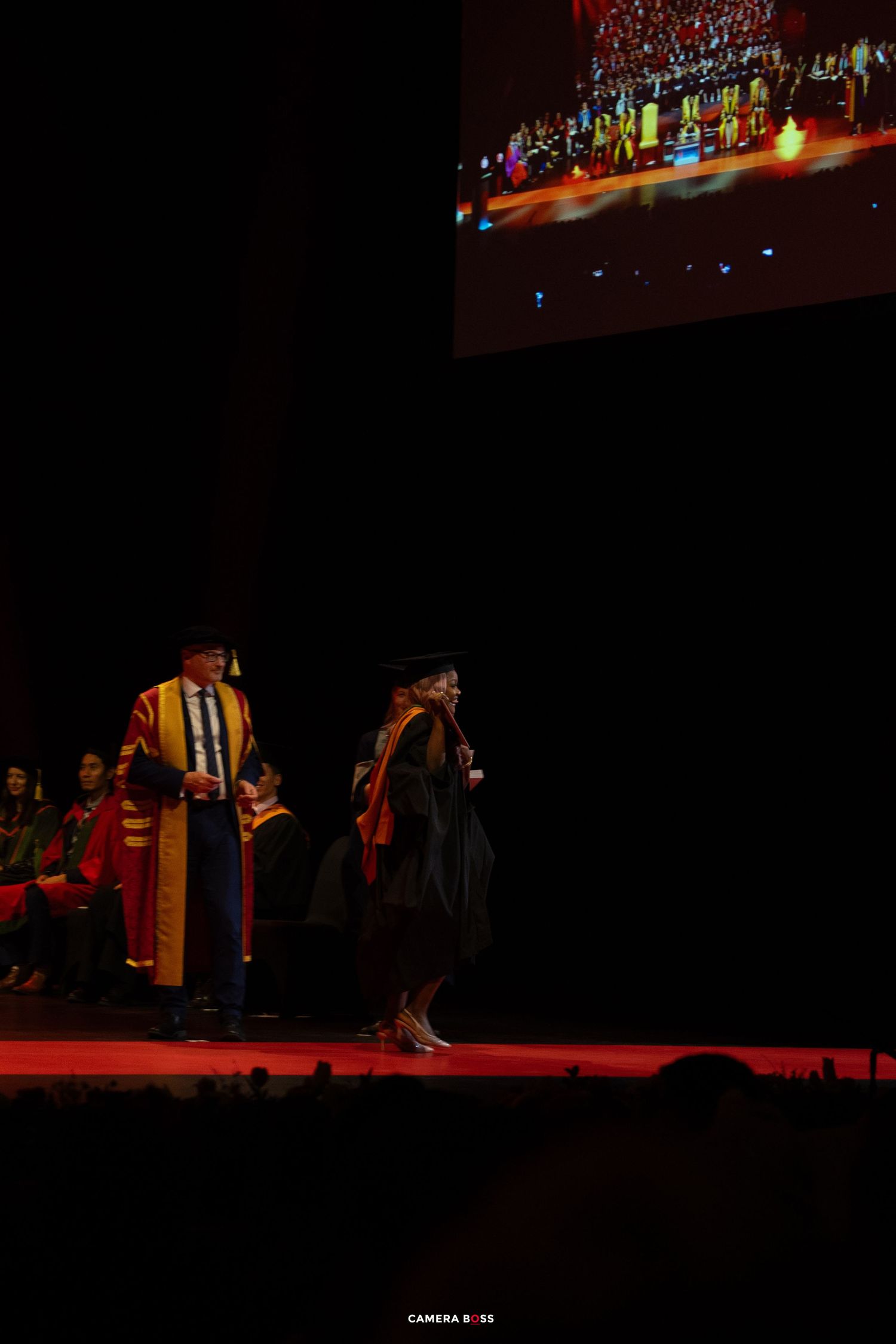 Graduates in academic regalia walk across a dimly lit stage during a commencement ceremony with red lighting effects.