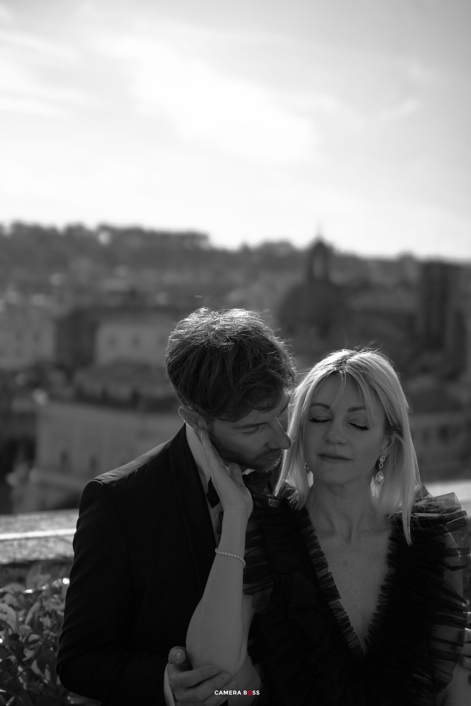 Romantic black and white photograph of a couple sharing an intimate moment on a rooftop overlooking a city landscape.