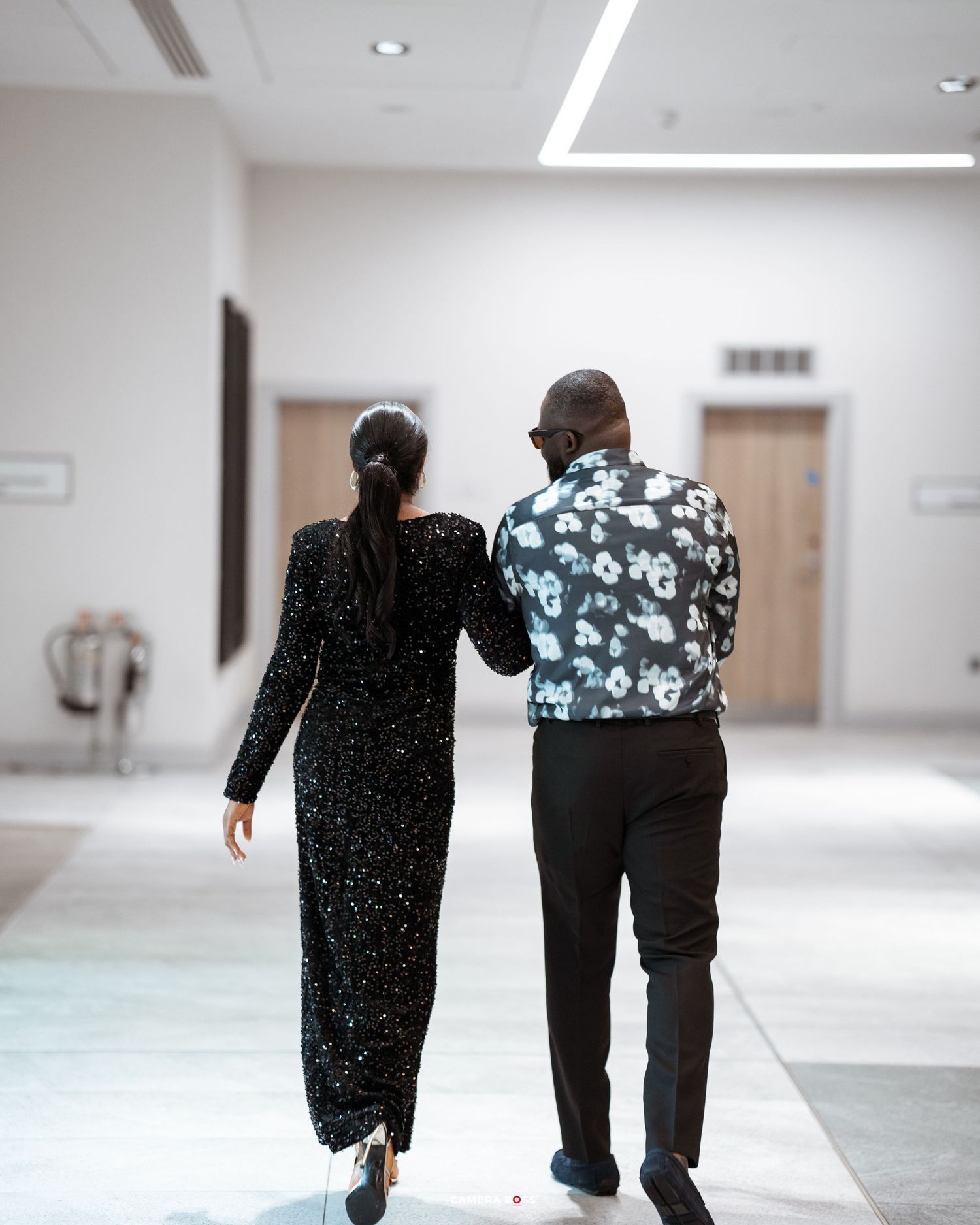 Two individuals in formal attire walking together down a bright hallway, viewed from behind.