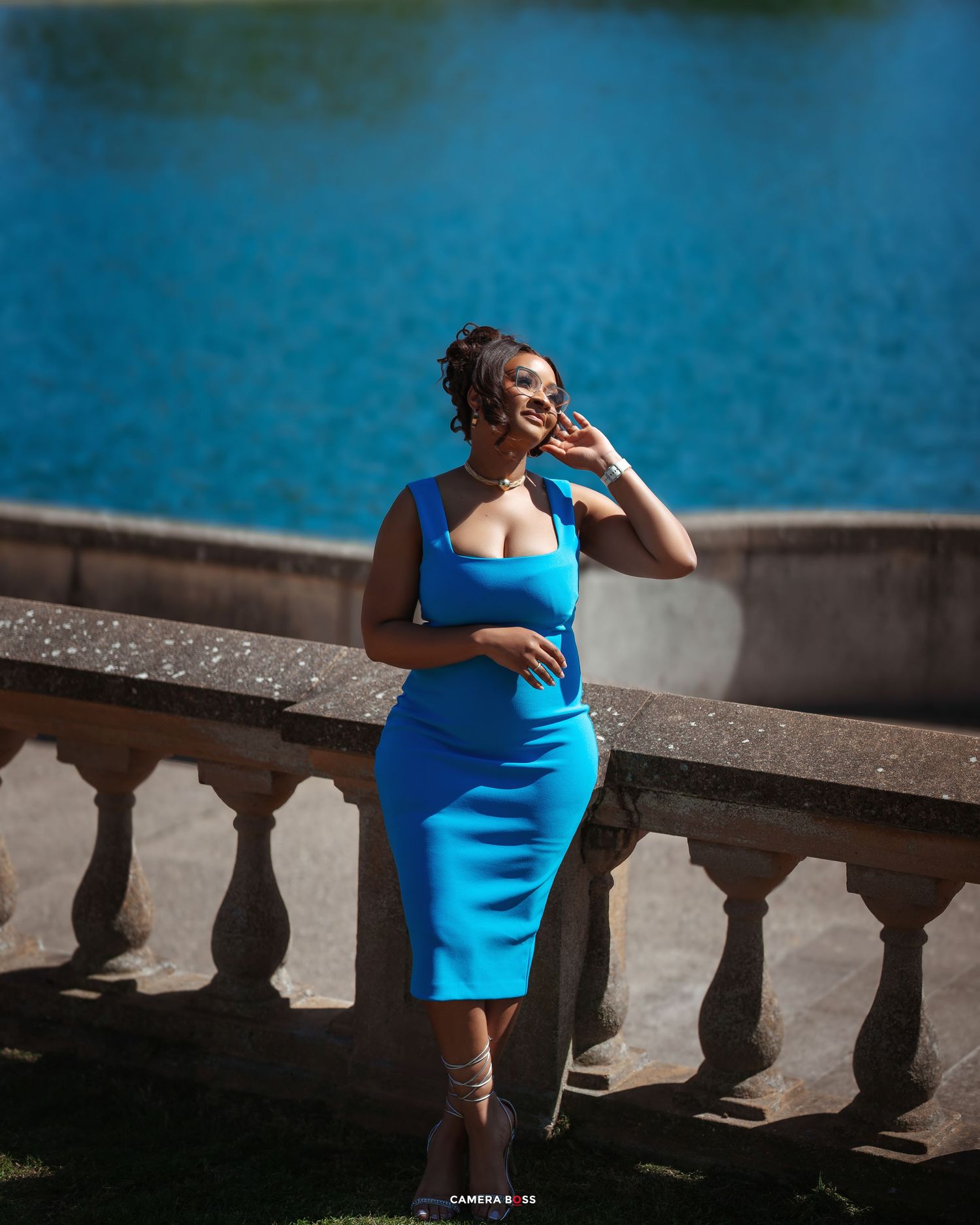 Person in a fitted bright blue dress poses elegantly against a stone balustrade with blue water in the background.