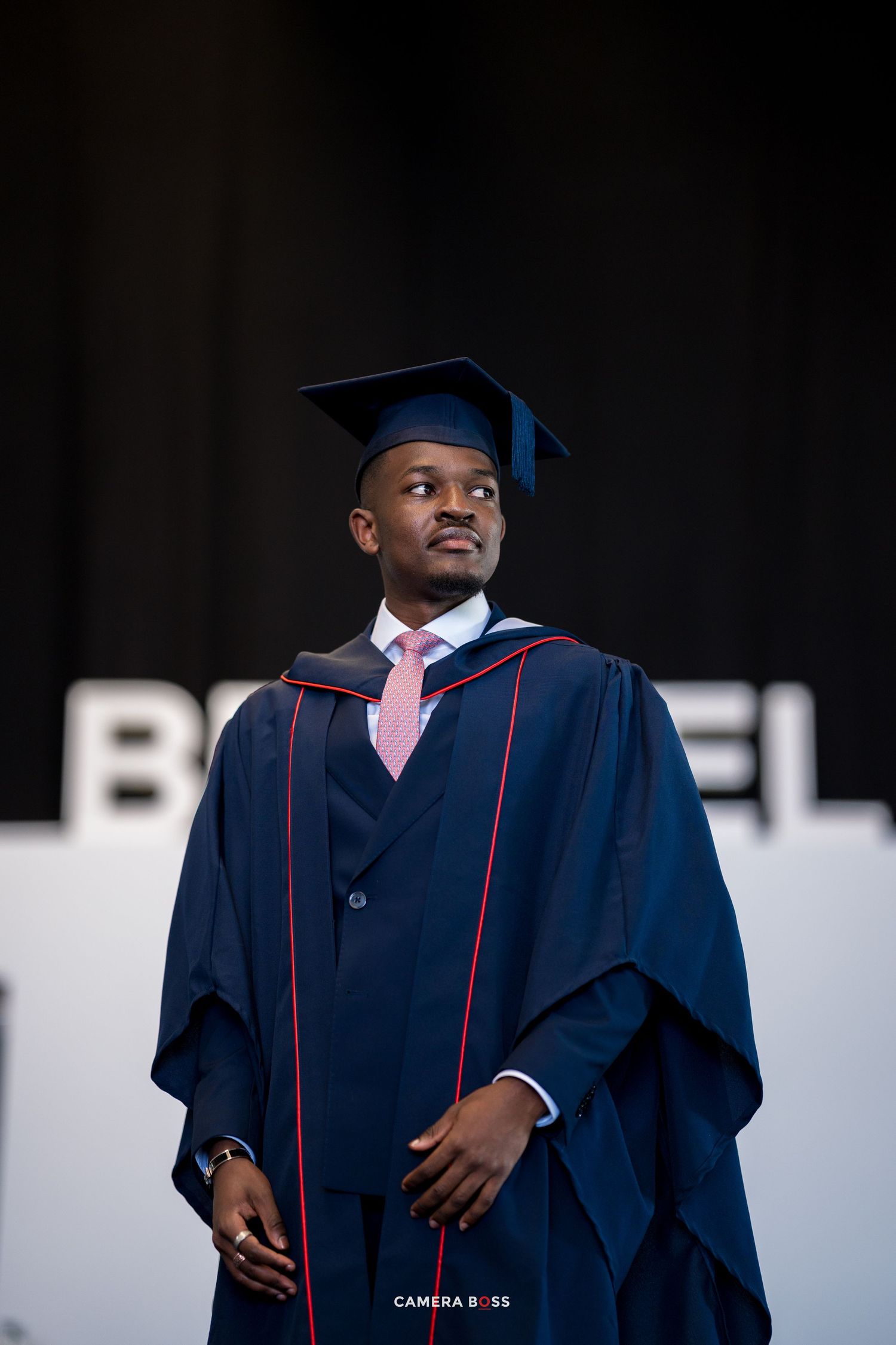 Graduate in dark blue academic regalia and pink tie stands against black backdrop during commencement ceremony.