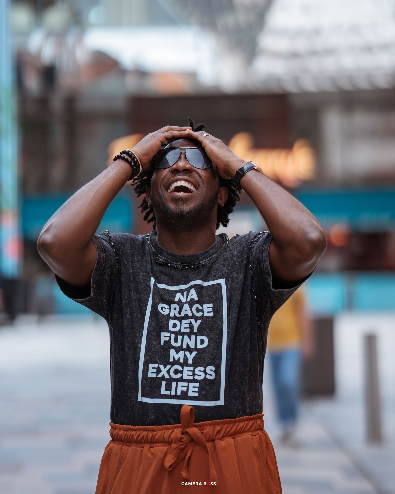 Person wearing a black graphic t-shirt and orange pants laughs joyfully on a city street.