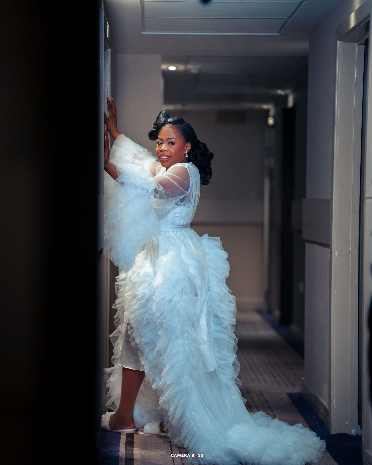 A person poses dramatically in a white ruffled tulle gown with a dramatic train in a hallway.