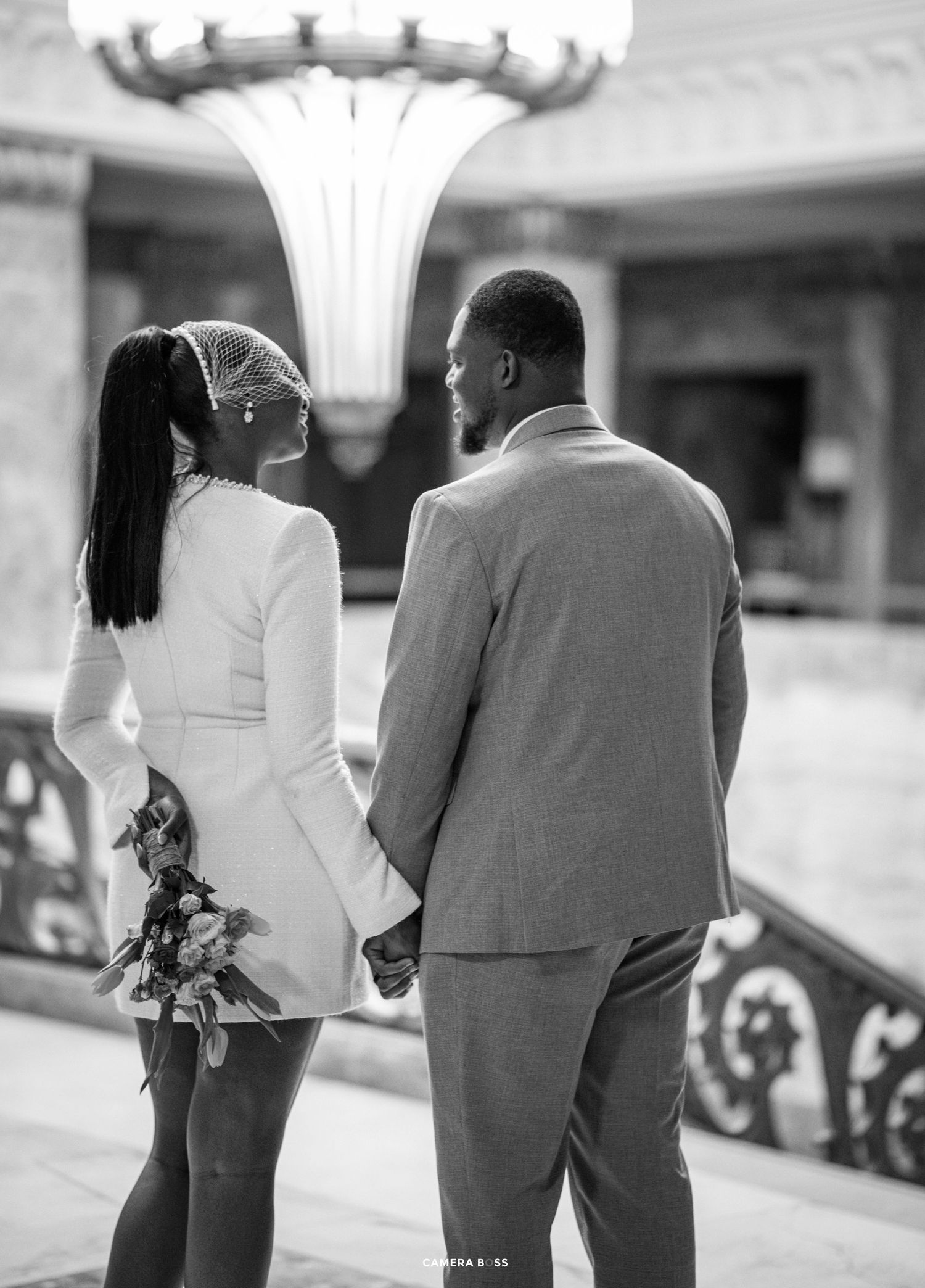A romantic black and white photo of a couple in formal attire holding hands while standing under an ornate light fixture.