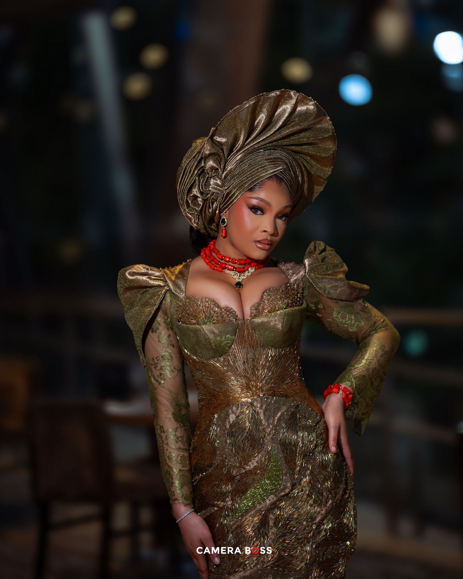 Elegant model in an ornate gold and green brocade gown with dramatic headpiece and red choker poses in dramatic lighting.