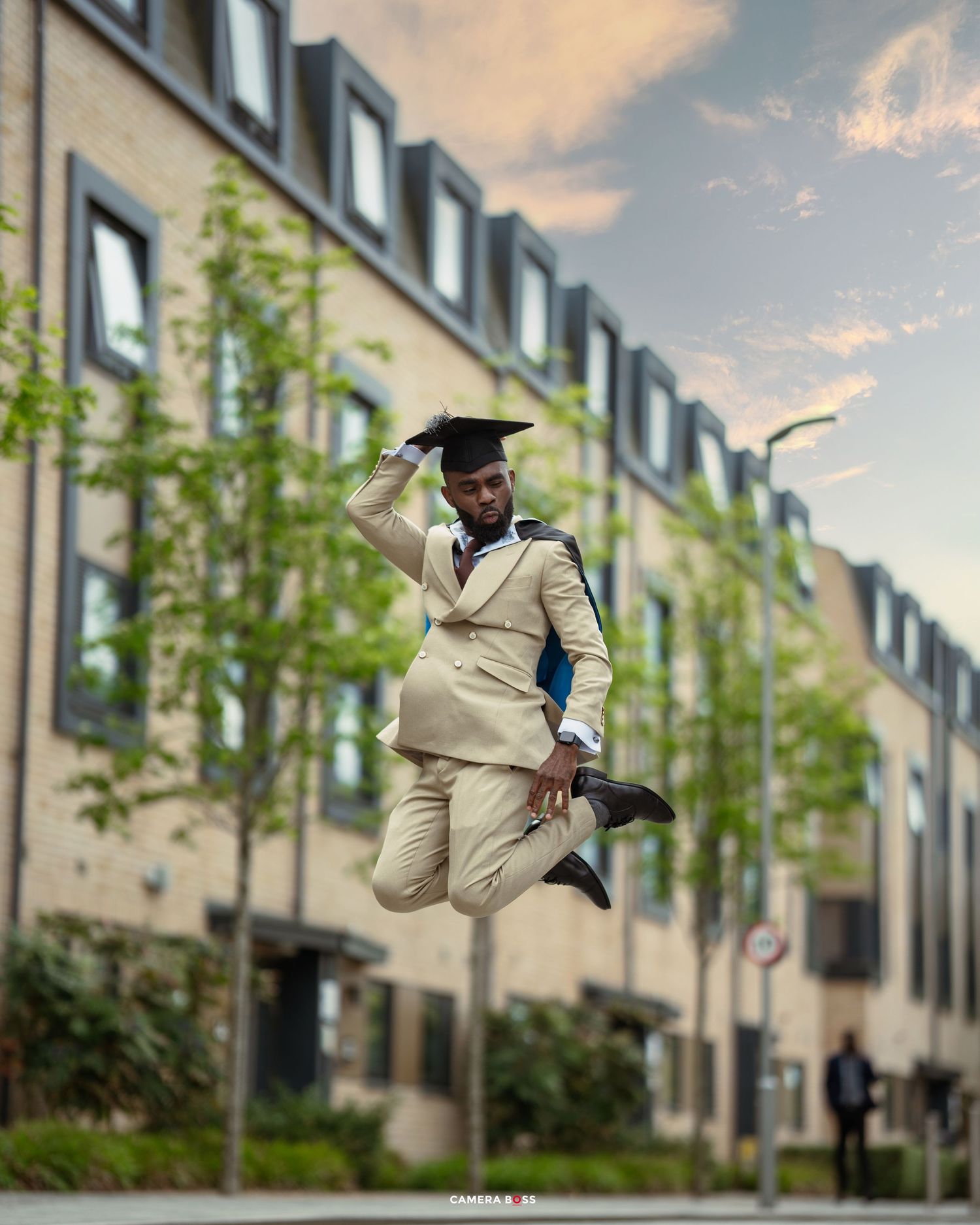 Graduate in a light colored suit and cap jumps joyfully in front of a modern apartment building on graduation day.
