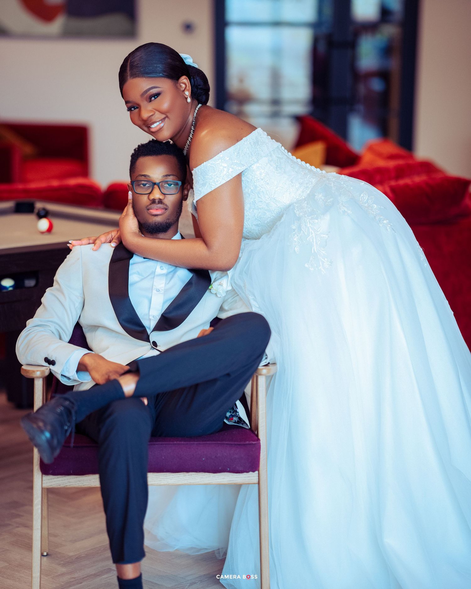 A couple poses intimately in formal attire, with the bride in a white off-shoulder gown embracing her partner on a purple chair.